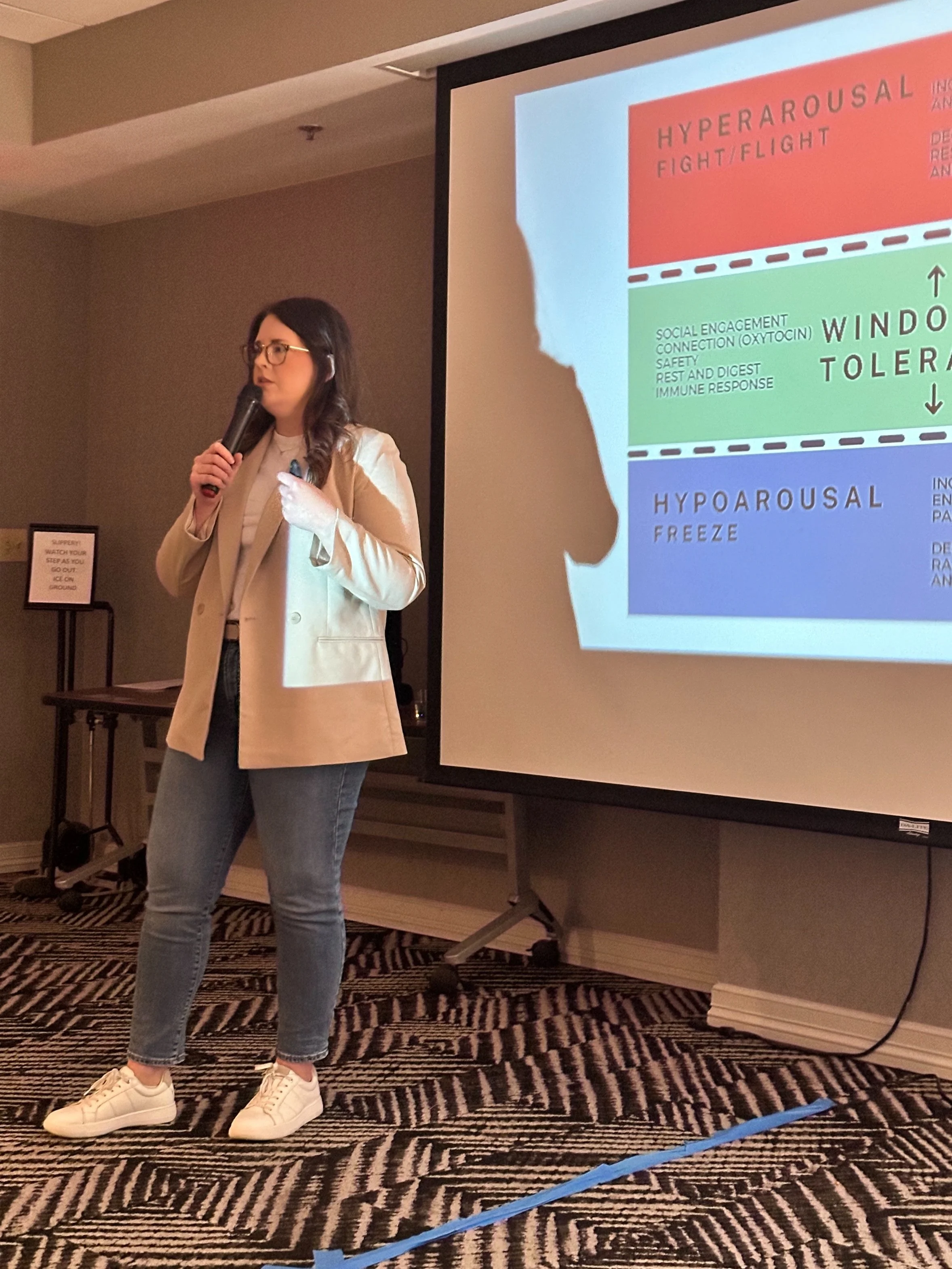 A woman holding a microphone giving a presentation in front of a slide titled "Hyperarousal Fight/Flight" with sections on window tolerance, hypoarousal freeze, and hyperarousal fight/flight.