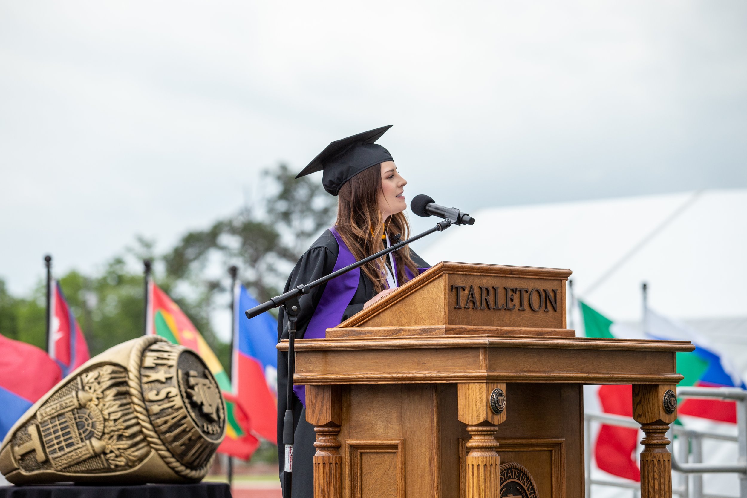Female graduate in a cap and gown speaking at a podium labeled 'Tarleton' during a graduation ceremony. Flags are visible in the background.