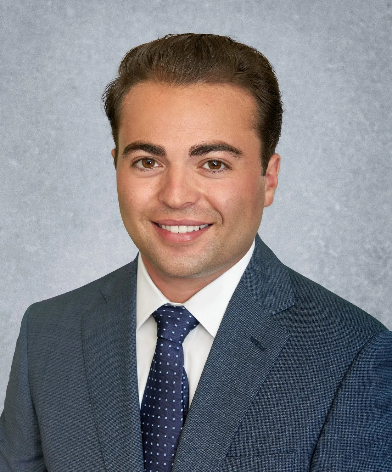 A professional headshot of a young man in a dark blue suit and tie, smiling, against a gray background.