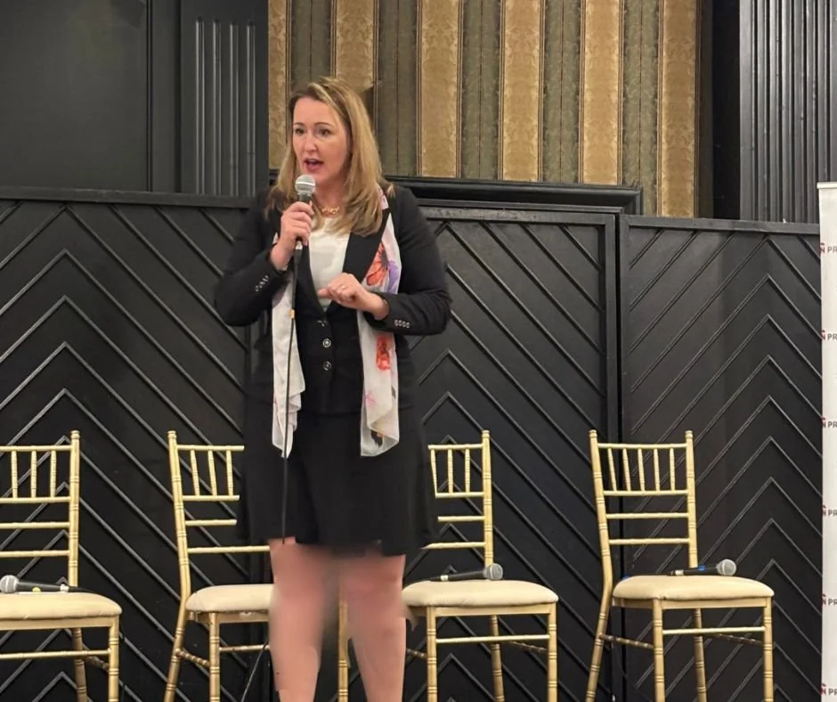 Kristina Holle-speaker in professional attire speaking into a microphone on stage with gold chairs behind her.