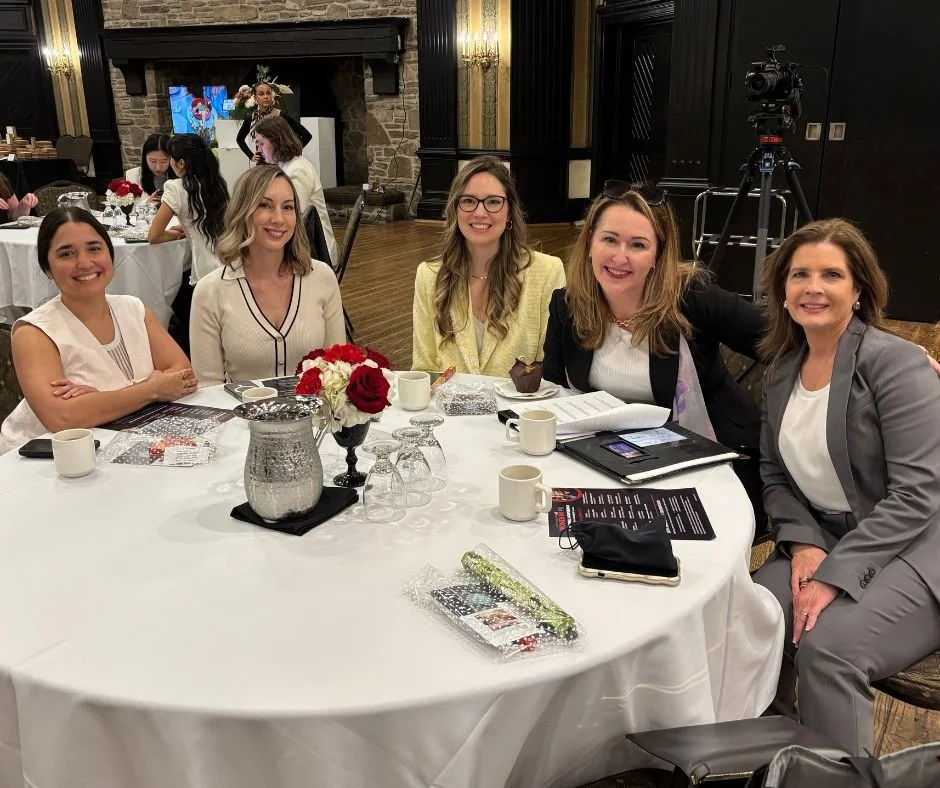 Kristina Holle with Group of five women sitting at a round table in a banquet hall Women in PR event , smiling at the camera, with others in the background, and a camera on a tripod in the corner.