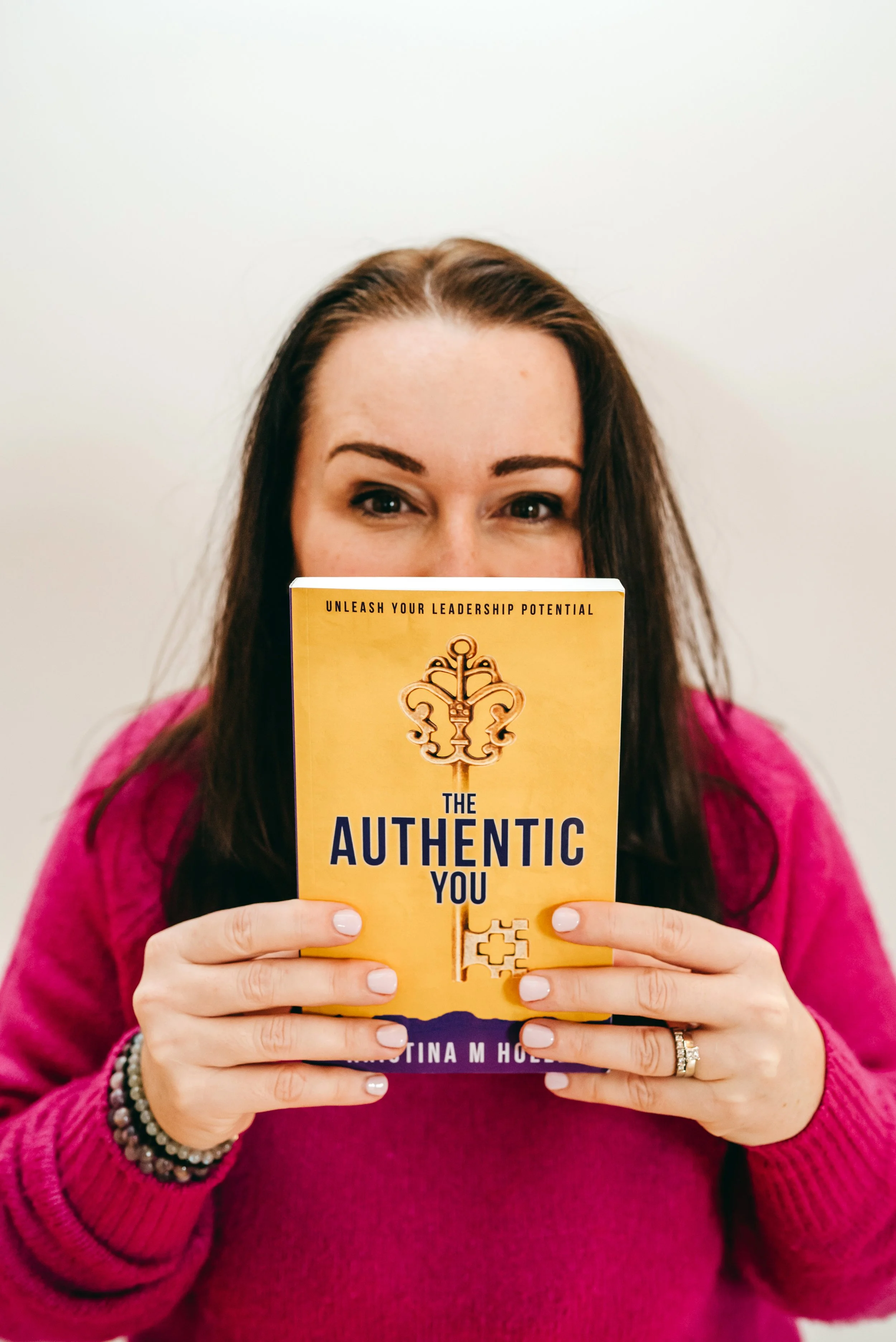 A person in a pink sweater holding a book titled "The Authentic You: Unleash Your Leadership Potential" by Tina M. Holmes.