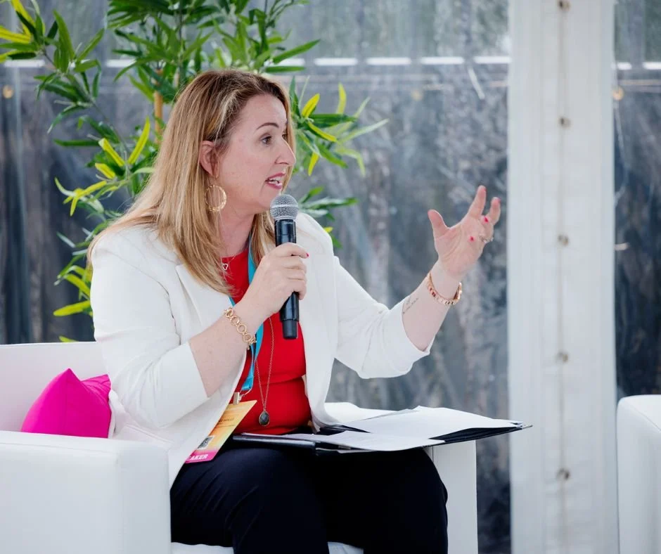 Kristina Holle speaking into a microphone while seated, with plants and a window in the background.