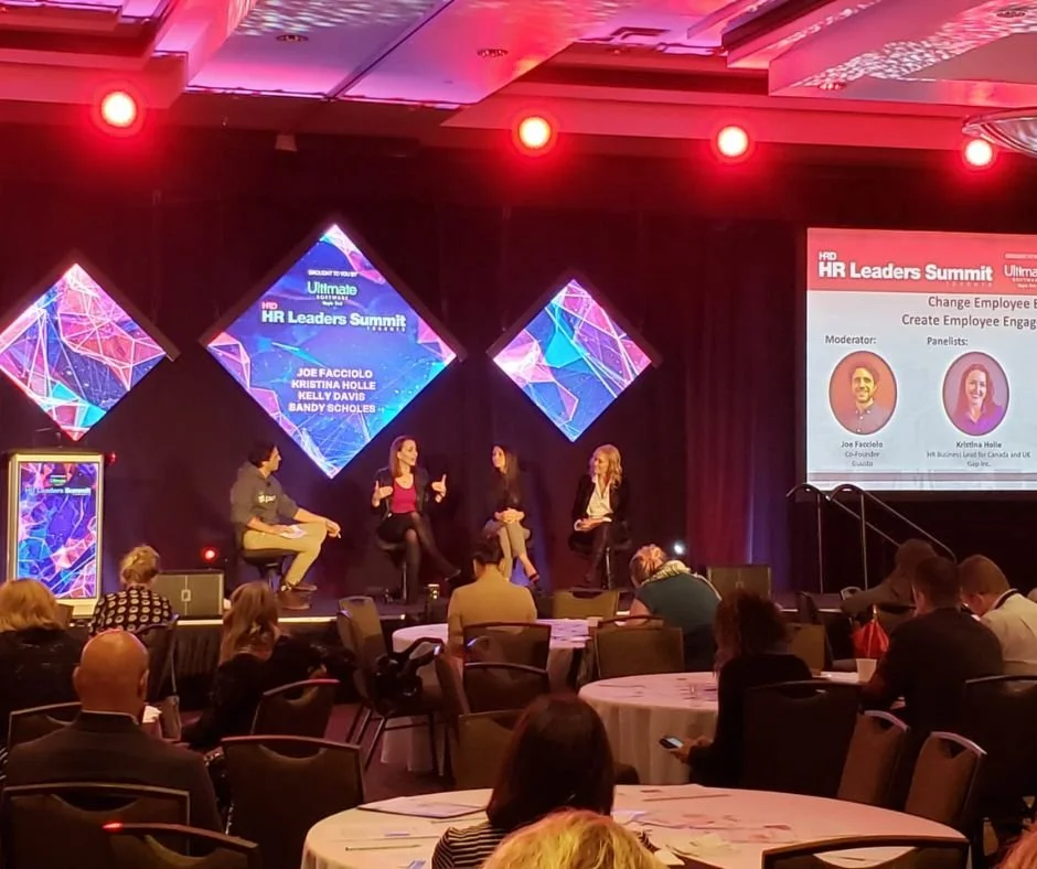 Krisitina Holle -speaker Panel discussion at HR Leaders Summit with three women and one man on stage, audience seated at round tables, large screens displaying event info and speaker names, stage illuminated with red and purple lighting.