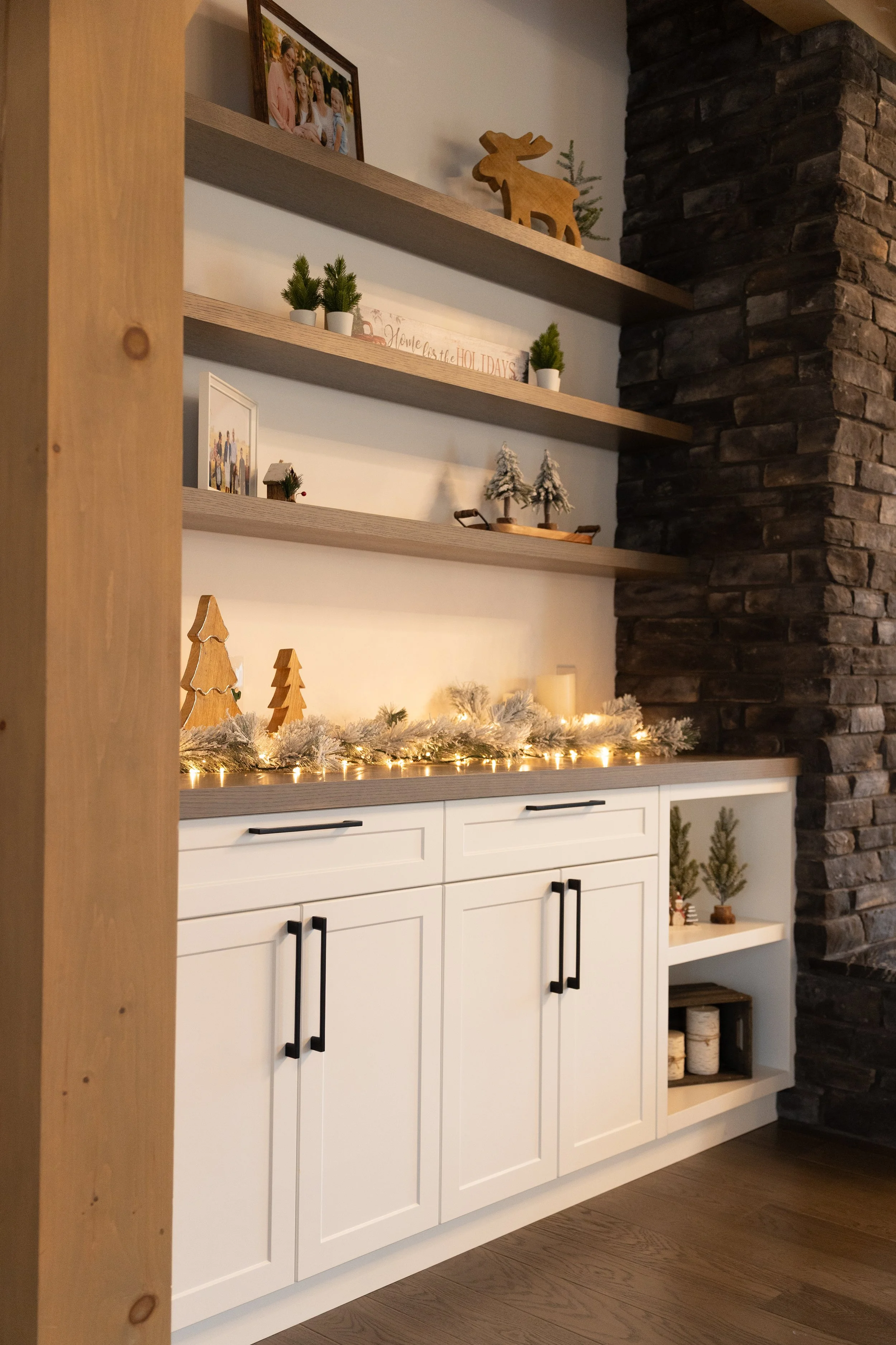 Close-up of a festive holiday decorated shelf with small Christmas trees, a garland with lights, candles, and decorative items. A brick wall is on the right, and a wooden picture frame is on top of the shelves.