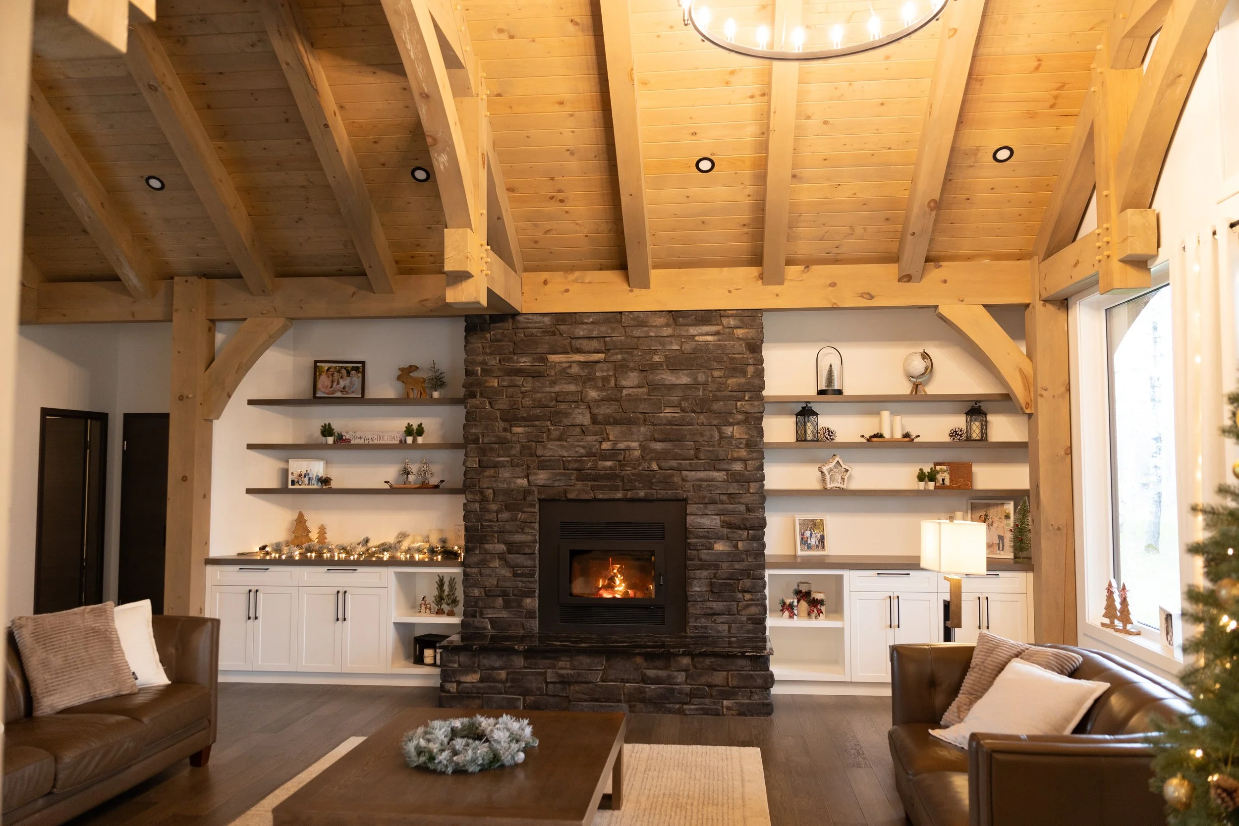 Living room with a stone fireplace, built-in white cabinets and open shelves decorated with holiday decor, a leather sofa, a coffee table with a green wreath, wooden beams on the ceiling, and a window with a Christmas tree nearby.