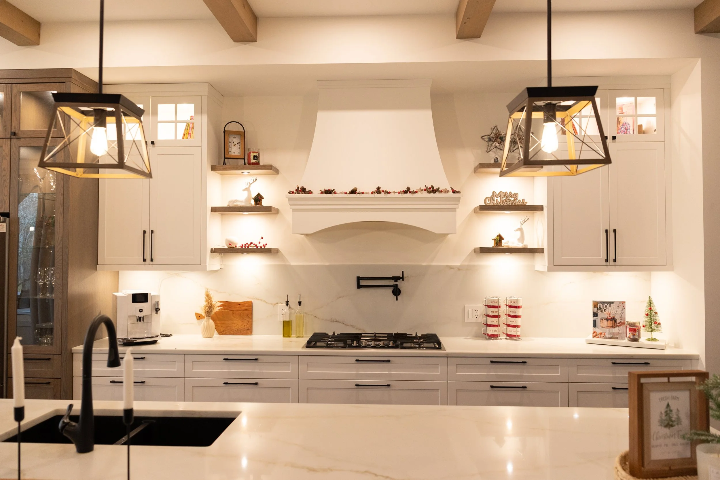 Modern kitchen decorated for Christmas with white cabinetry, a white range hood, open shelves with holiday decorations, and two geometric pendant lights.