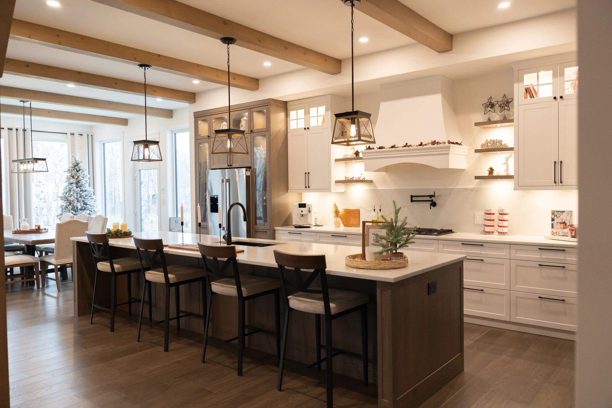 Modern kitchen with white cabinets, a large island with seating, pendant lights, and holiday decorations. Dining area with a Christmas tree visible through large windows.