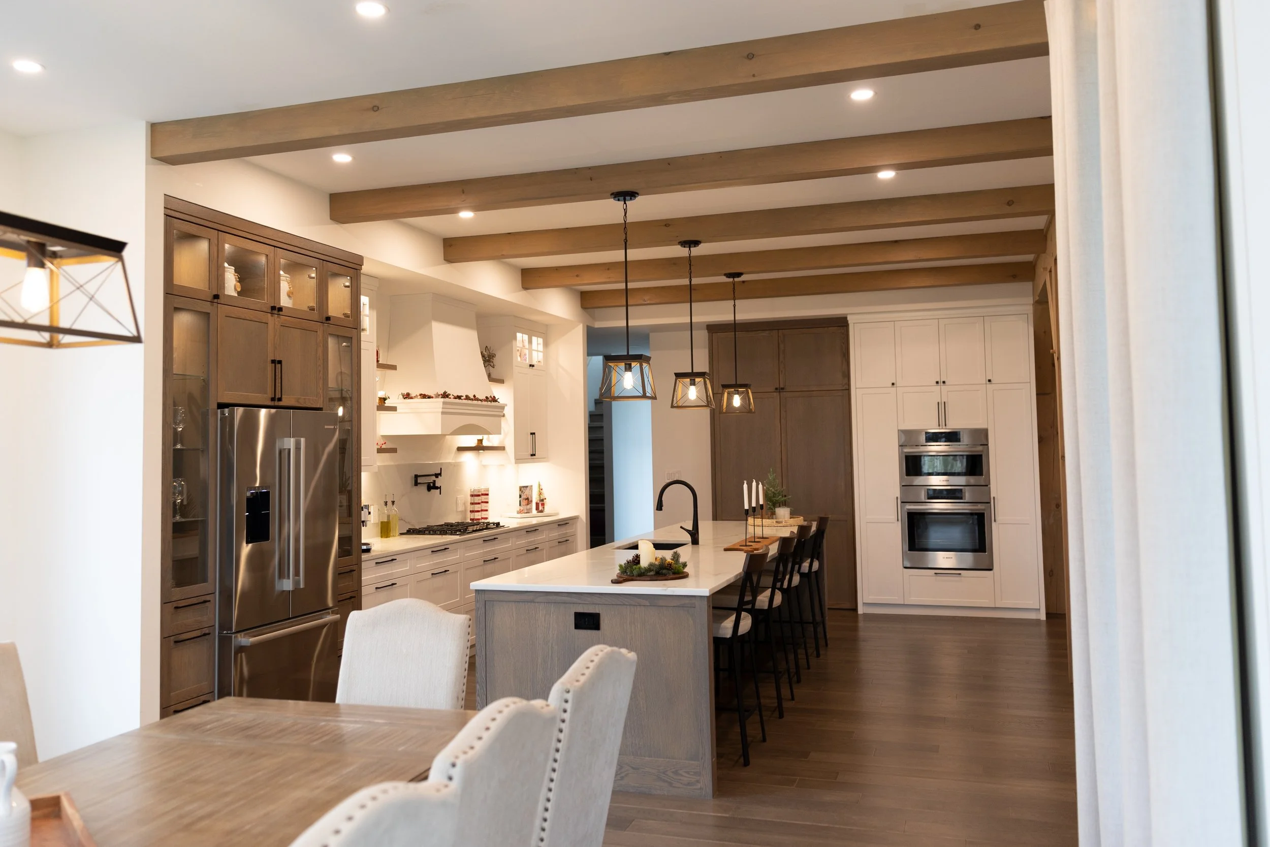 Modern kitchen with white cabinets, a large island with seating, wooden beams on the ceiling, stainless steel appliances, and pendant lighting.