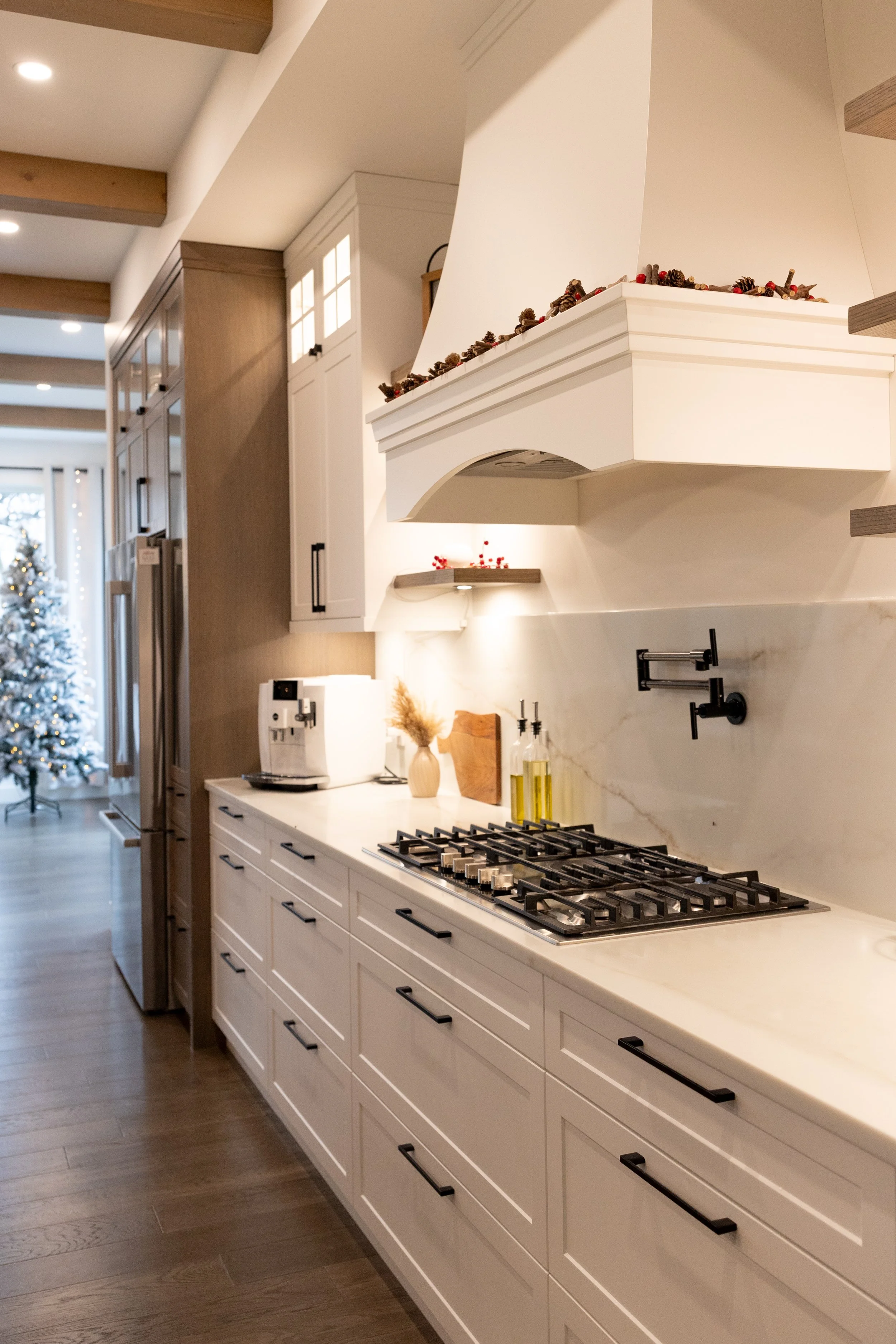 A modern kitchen with white cabinets, a marble backsplash, a gas stove, and Christmas decorations, including a Christmas tree in the background.