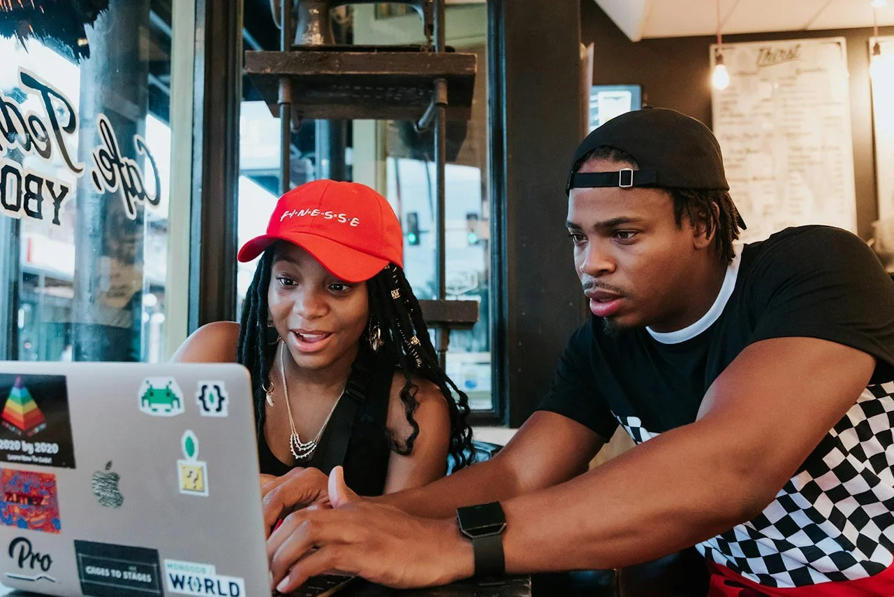 Two people sitting at a table using a laptop, one wearing a red cap and the other a black cap, in a casual setting. They are remotely building a life insurance agency that is protecting families and building generational wealth.