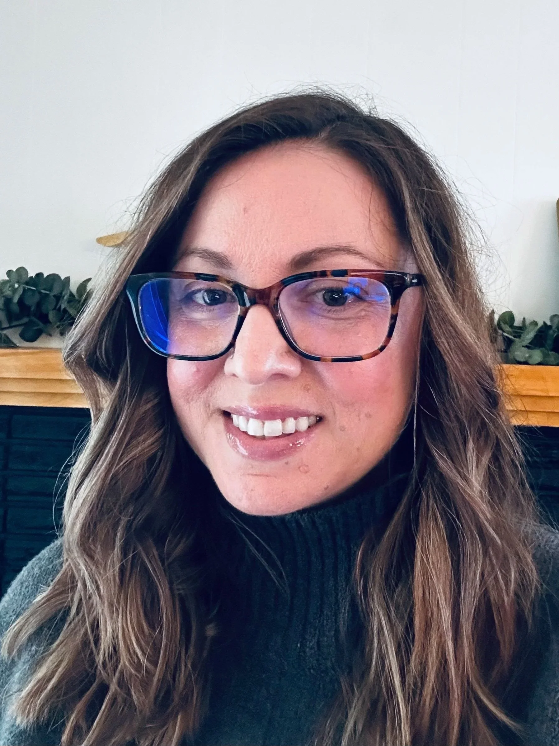 A woman with long wavy brown hair, glasses with tortoiseshell frames, and a black turtleneck sweater, smiling indoors with a white wall and decorative greenery in the background.
