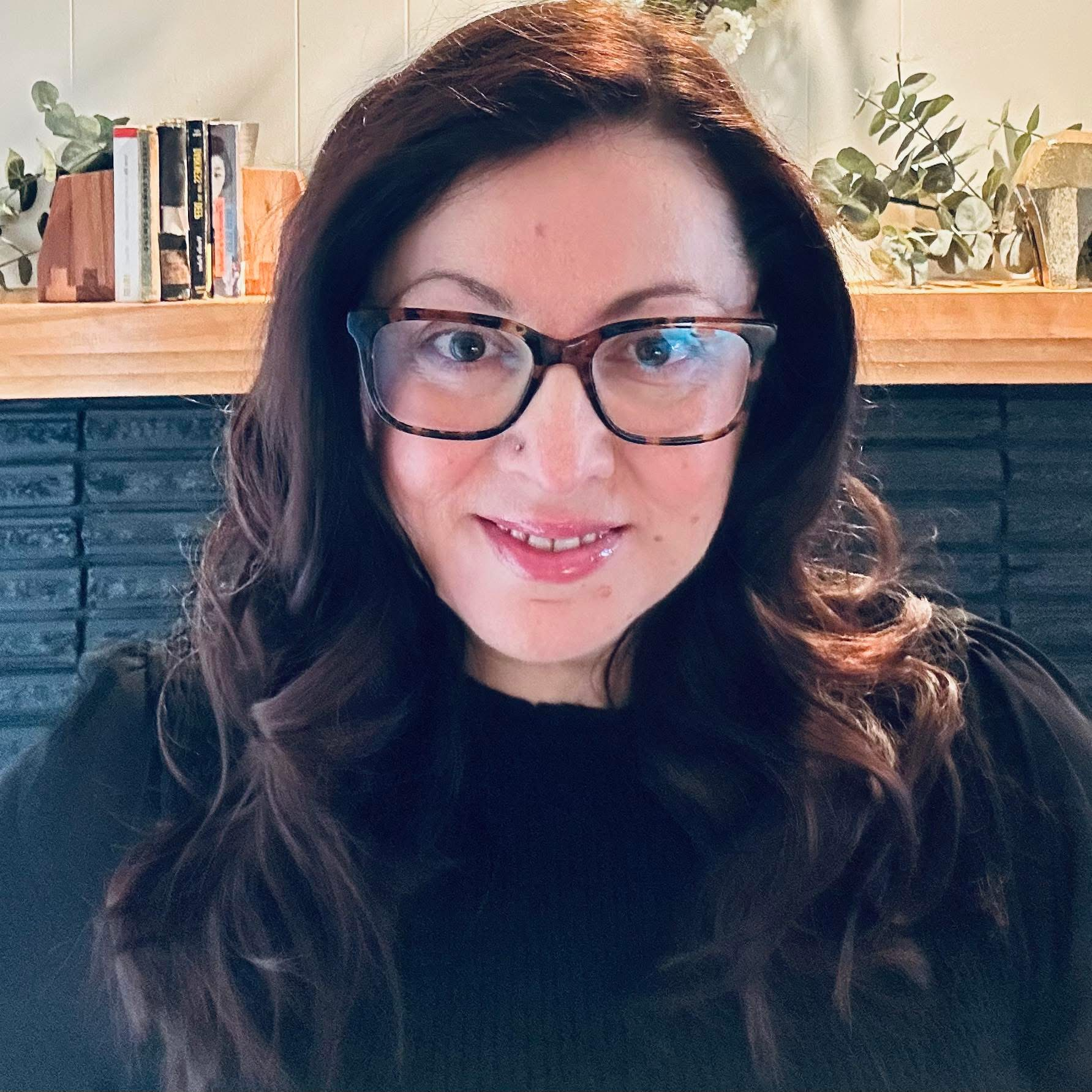 A woman with long wavy dark brown hair wearing glasses smiling in a cozy indoor setting with a brick fireplace, books, and decorative plants on a wooden shelf in the background.