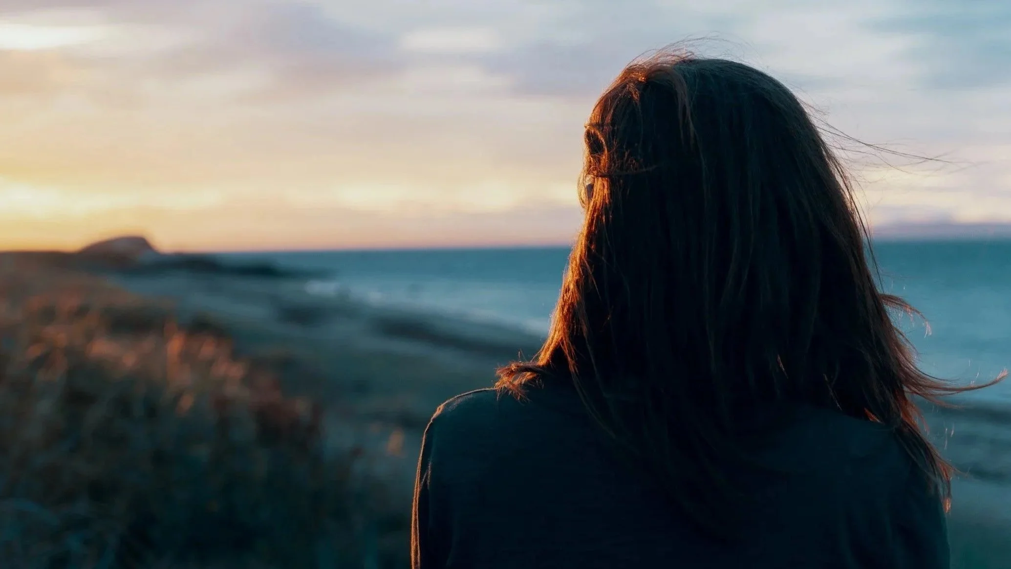 A woman with dark hair looking at the ocean during sunset, with the sky painted in soft pastel colors.