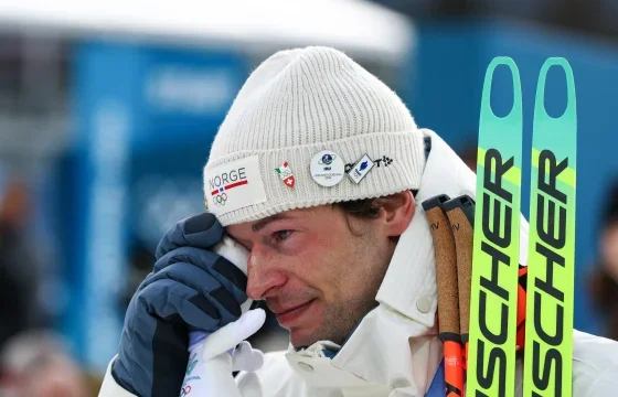 Close-up of a male skier wearing a white knit hat with Olympic pins, holding ski poles, with a blurred background.