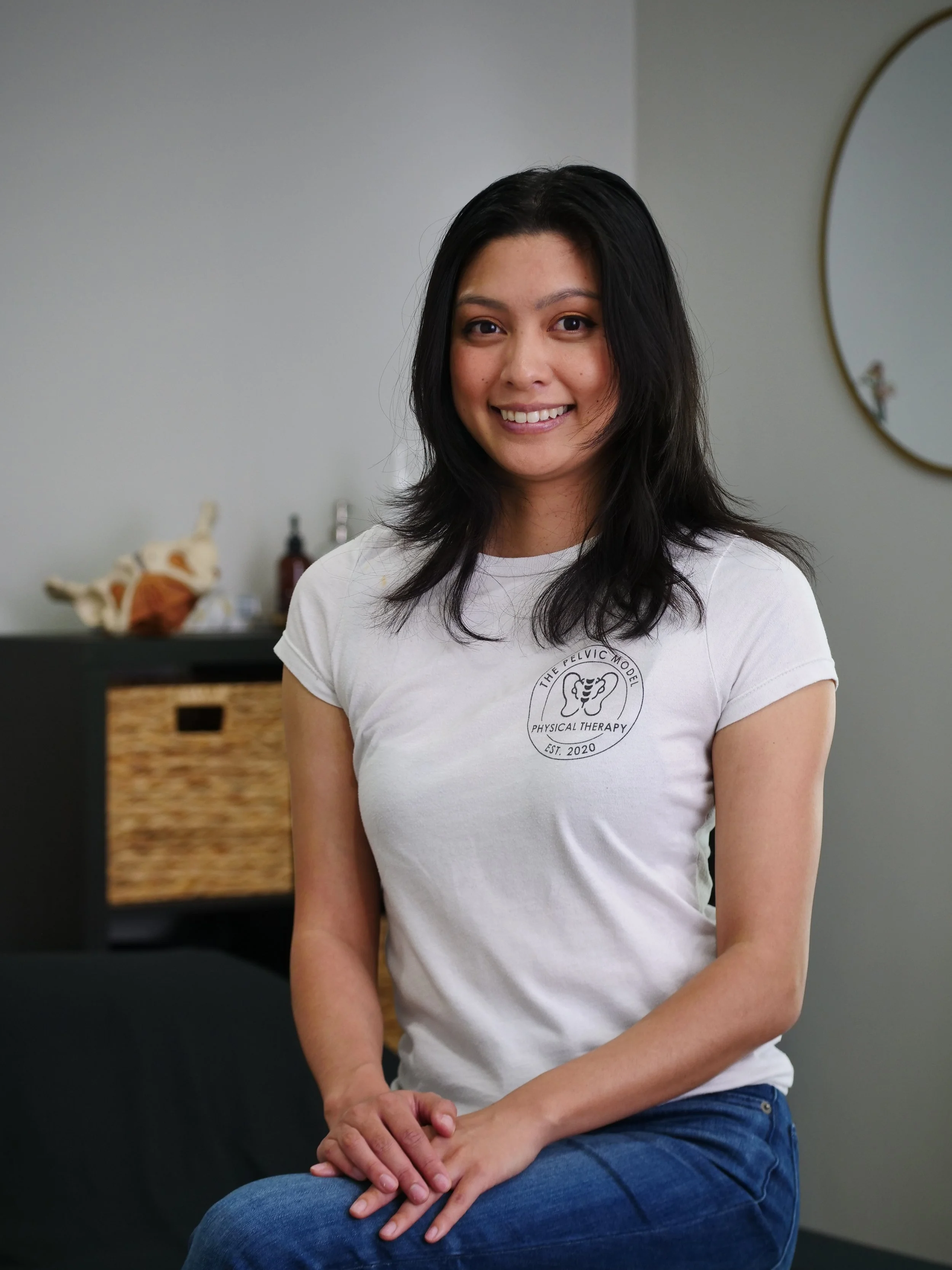A woman with black hair smiling, wearing a white T-shirt with a logo for physical therapy, sitting with her legs crossed in a room with a mirror and a basket in the background.