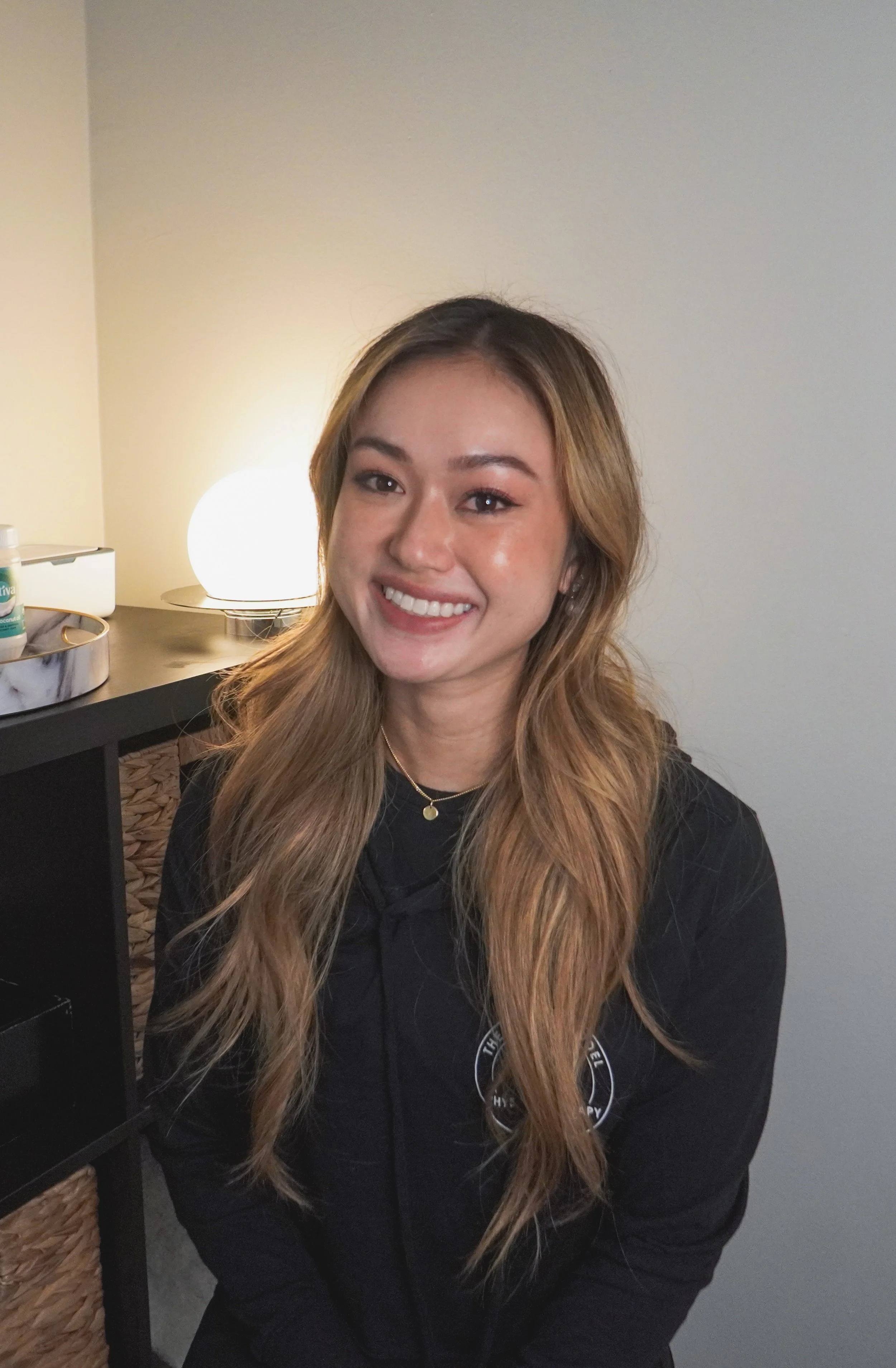 A young woman with long wavy hair smiling while sitting indoors. She is wearing a black hoodie with a logo on it and a gold necklace. In the background, there is a lamp and some items on a black piece of furniture.