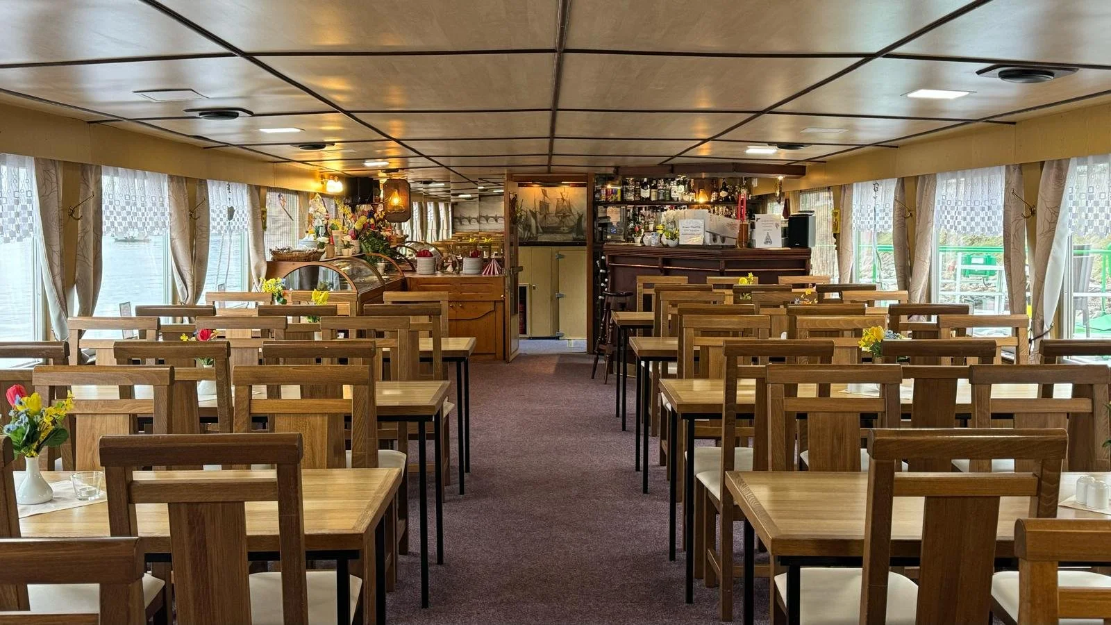 Interior of a dining area on a boat with wooden tables and chairs, decorated with flowers, and a view of the bar at the back.