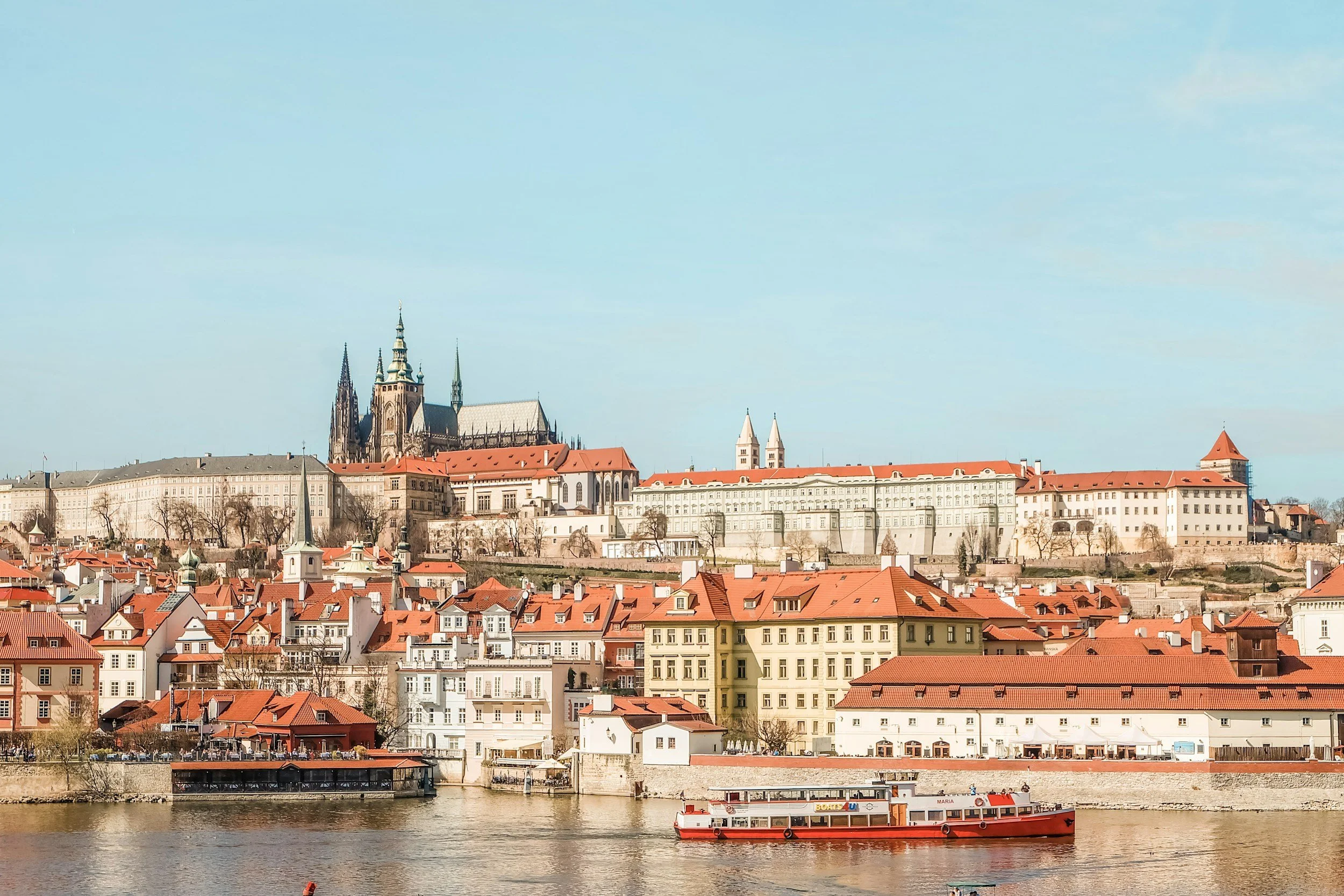 View of Prague Castle and Vltava River with red-roofed buildings and a boat in the foreground.