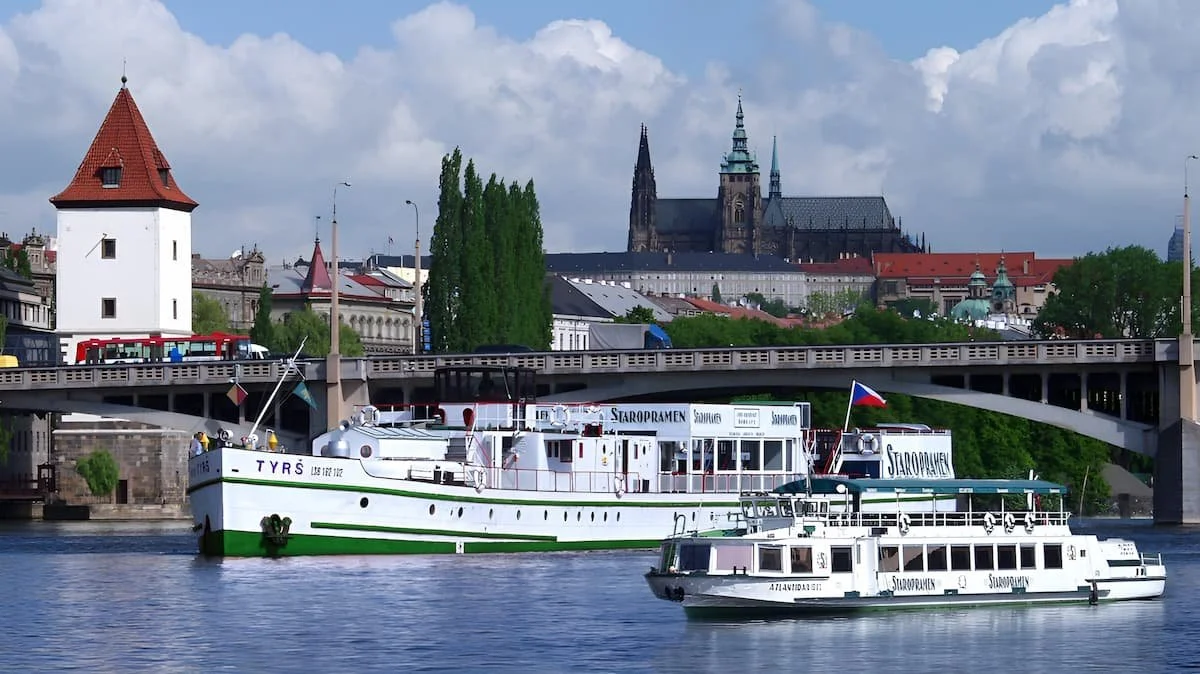 Two private boats on a river with a cityscape and historic buildings, including a large cathedral, in Prague