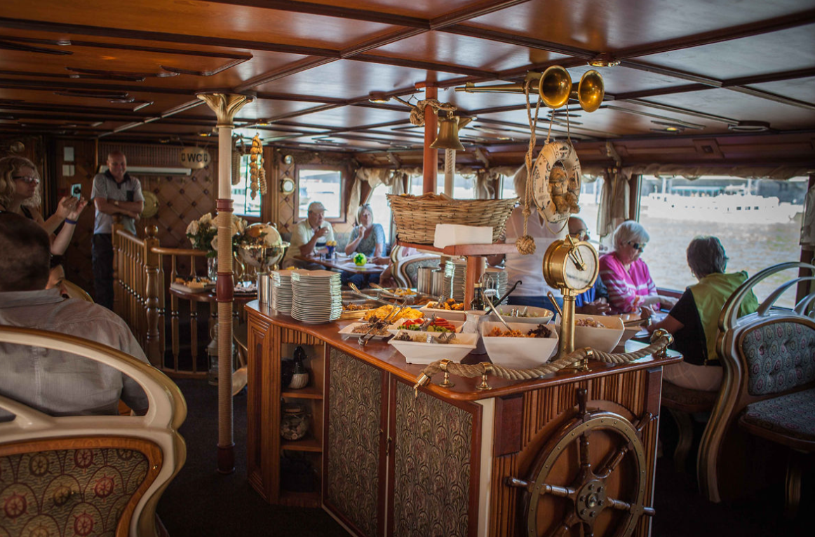 Interior of a dining area on a boat with people seated by large windows looking outside at water. A buffet table with plates, bowls of food, and decorative nautical items is in the center.
