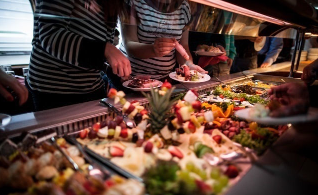 People serving themselves food from a buffet with assorted desserts, fruits, and salads.