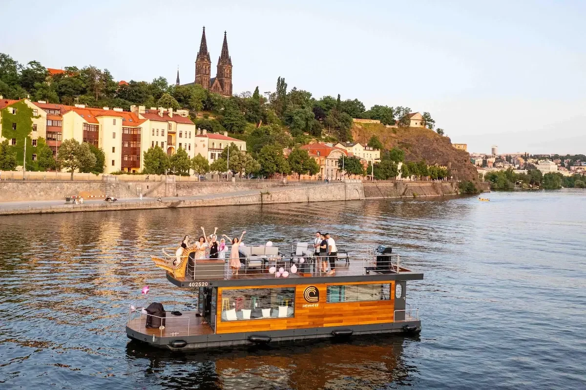 A rental boat with people on the upper deck celebrating, with a Prague city and church steeples in the background.