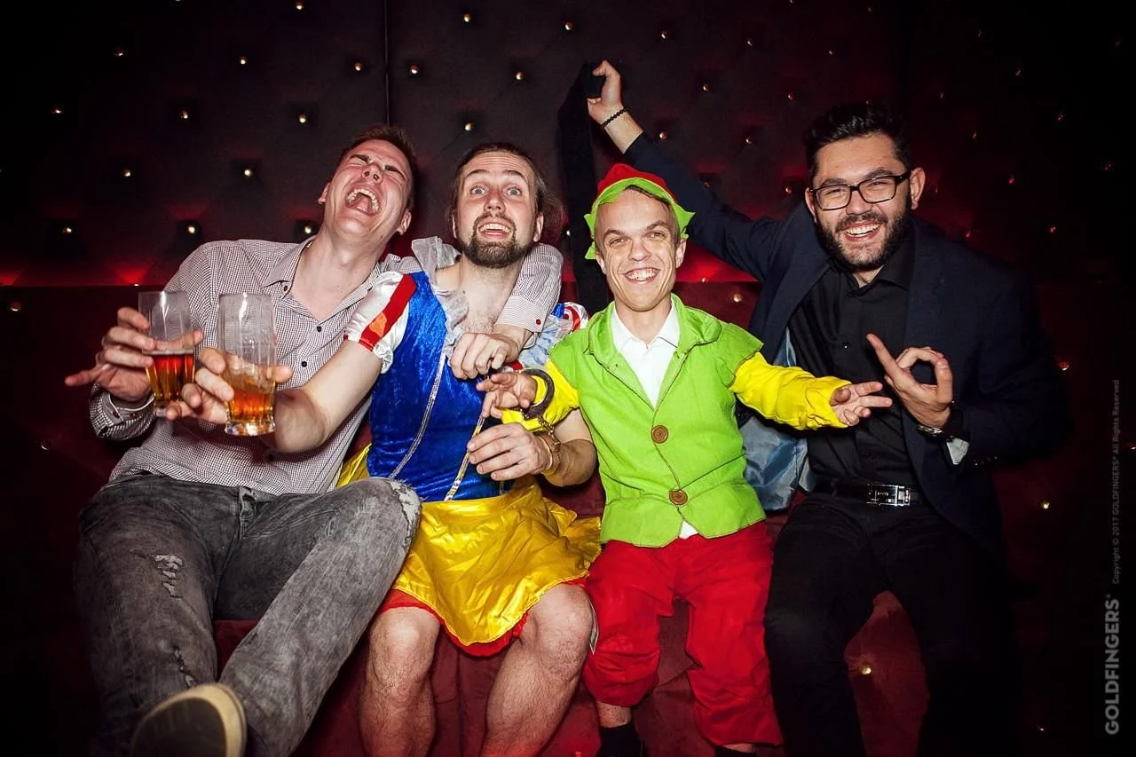 Group of men celebrating a bachelor party, one dressed as Snow White, another as a dwarf, laughing and holding drinks