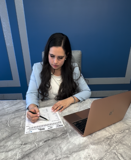 Woman sitting at a marble table, working on documents with a pen, with a laptop open in front of her in a blue office or conference room.