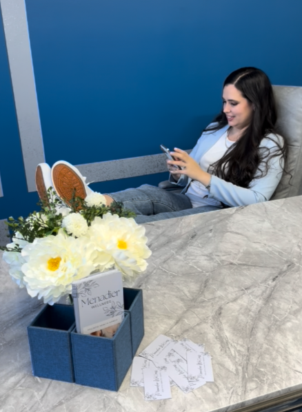 A woman with long dark hair sitting at a marble table, smiling and looking at her phone. On the table, there is a blue container with white flowers and some papers, and in the background, a blue wall.