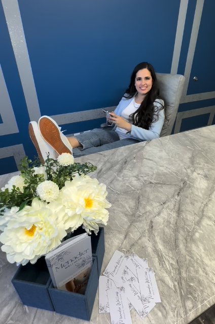 Woman sitting in an office chair at a marble table, smiling, with feet resting on the table, holding a phone. A vase of white flowers and some papers are on the table, and the background features blue walls with decorative gray stripes.