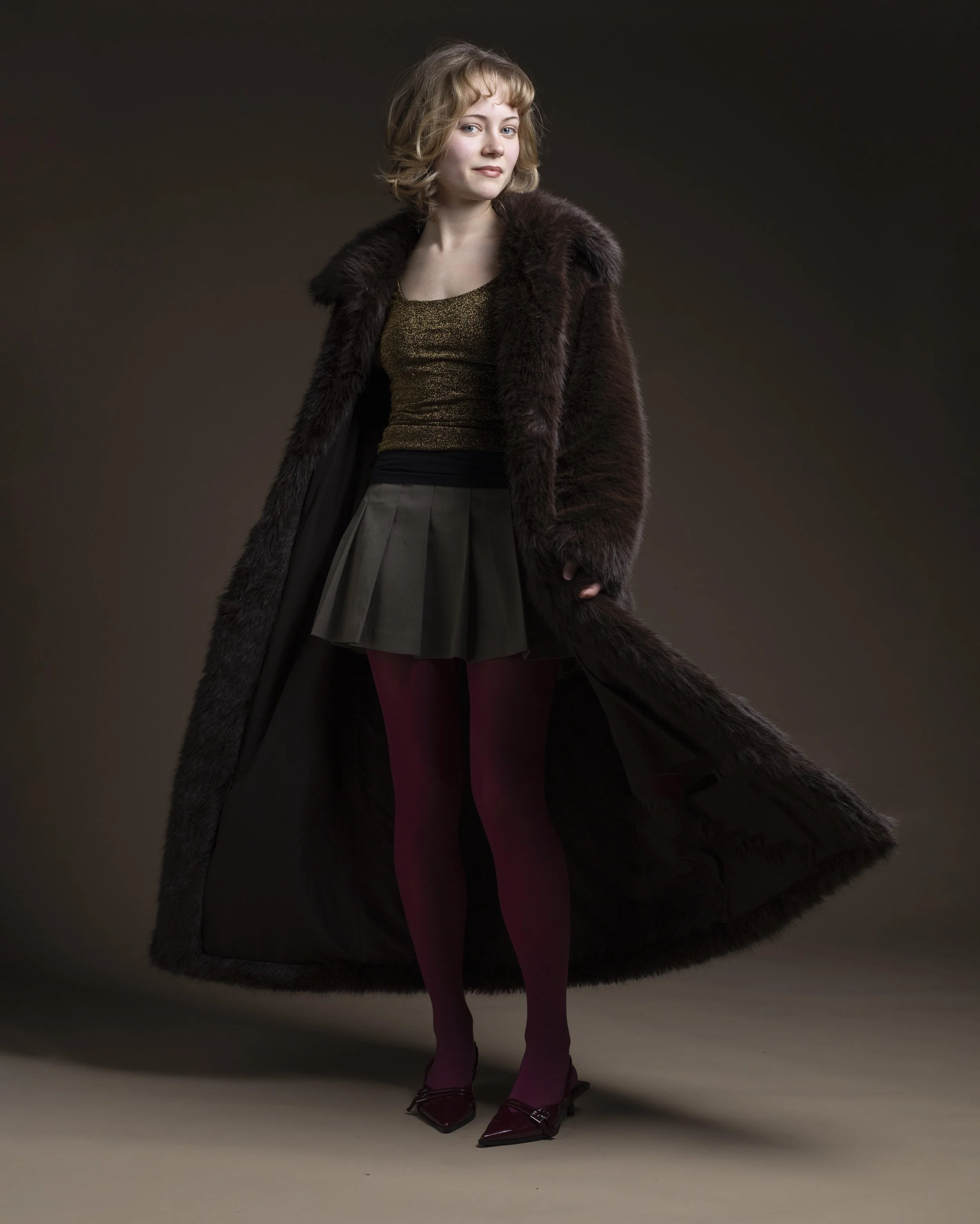 En kvinne står mot en mørk bakgrunn, iført en lang pelskåpe, en gullfarget topp, en svart skinnskjørt, vinrøde strømper og høye hæler.