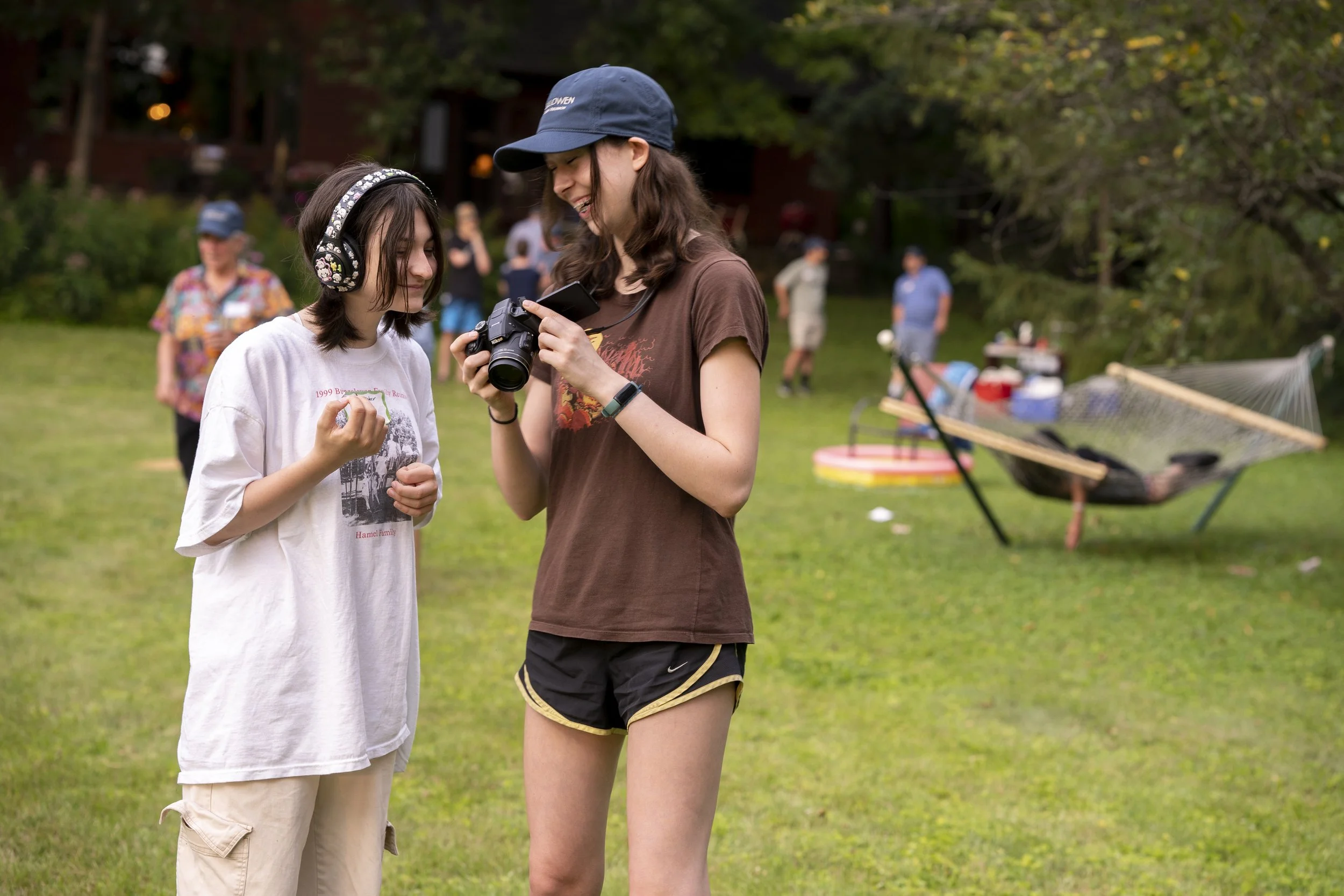 Two young women are standing on the grass at an outdoor gathering. One woman, wearing headphones and a large t-shirt, is looking at her hand while the other woman, wearing a cap and a brown t-shirt, is showing her something on her camera. In the back
