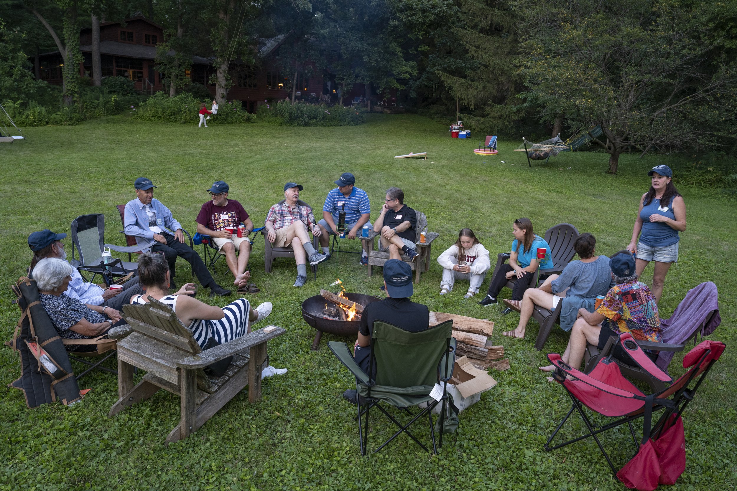 A group of people gathering in a backyard around a fire pit, sitting in chairs and talking, with some holding drinks. The scene is set in the evening with trees and a house in the background.