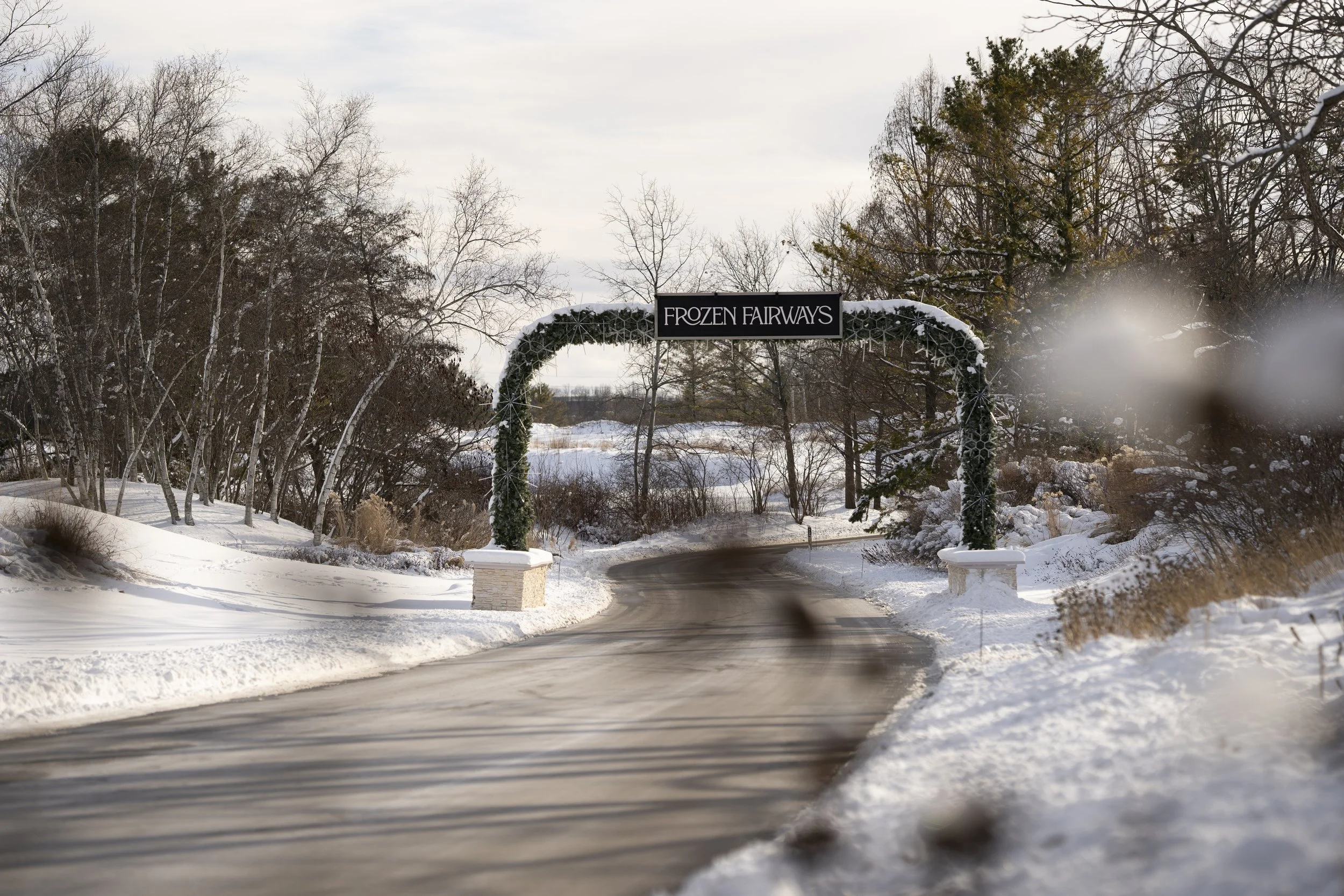 Snow-covered entrance to Frozen Fairways golf course with an archway and sign, trees, and a winding road in winter.