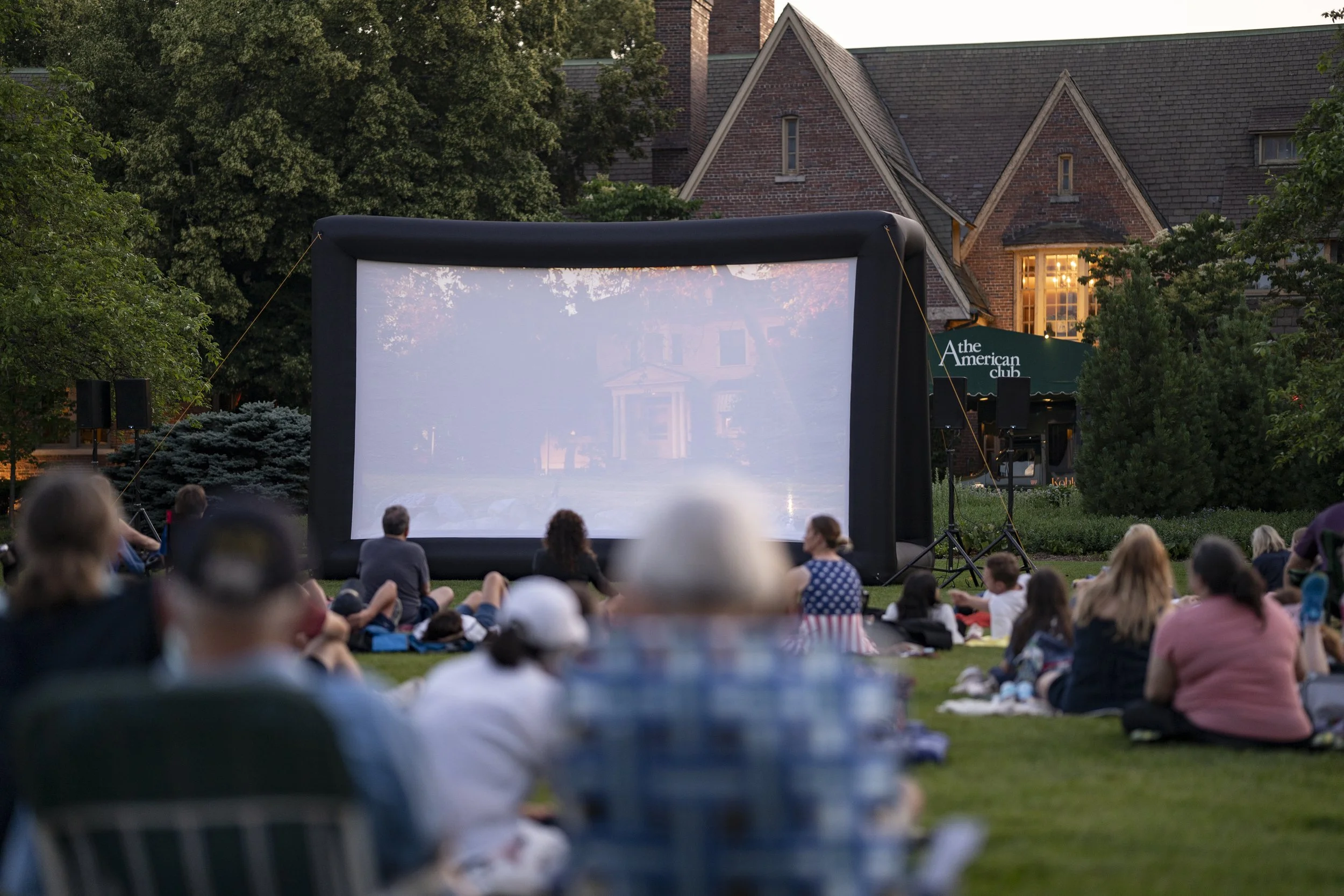 People sitting on a lawn watching an outdoor movie projected onto a large inflatable screen in front of a house with trees and a sign that says 'The American Club'.