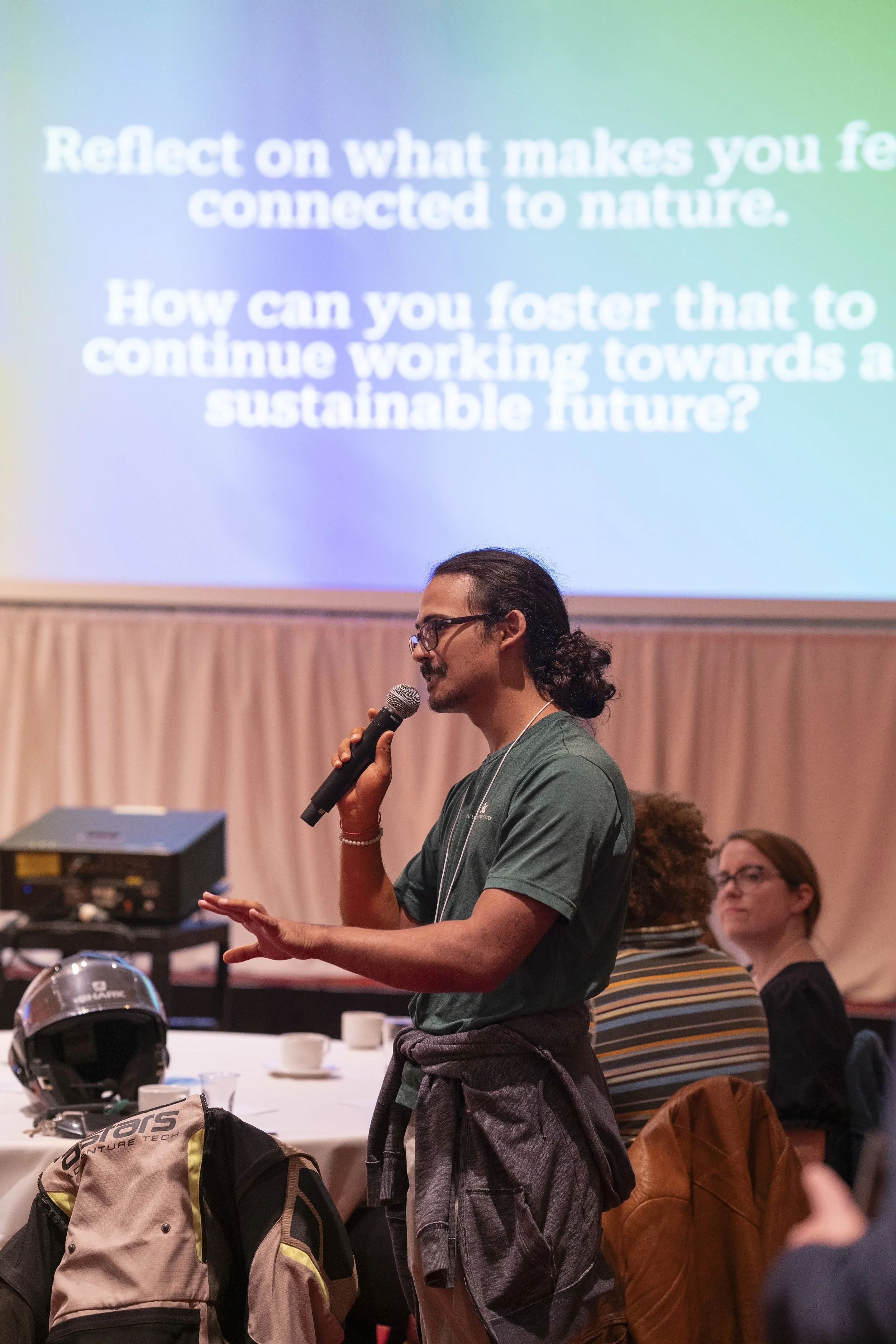 A speaker with long dark hair, beard, glasses, wearing a green t-shirt, is holding a microphone and giving a presentation in front of a large screen that displays a message about reconnecting with nature and sustainability. Several people are seated 