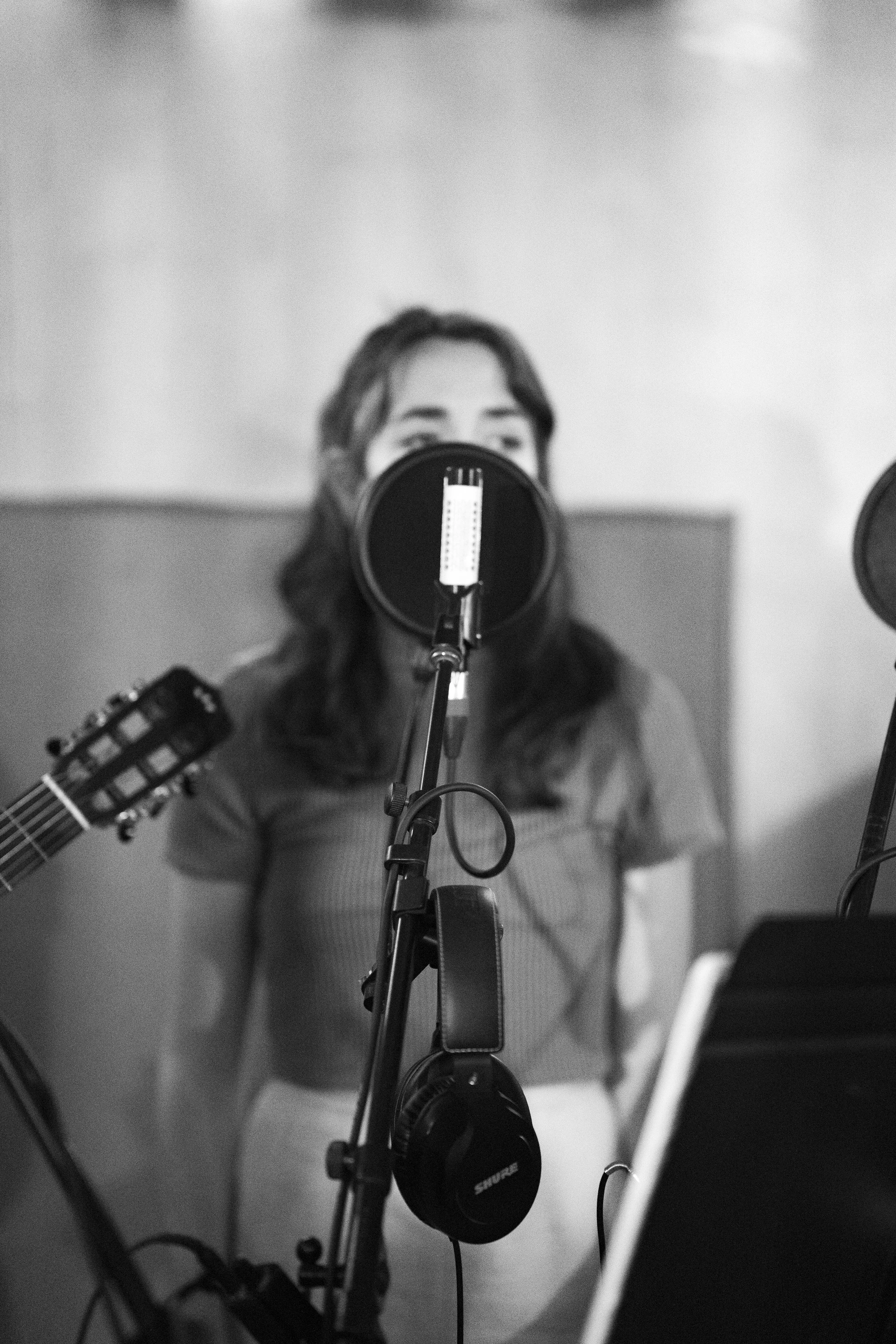 A woman singing into a microphone in a recording studio, with a guitar visible on her left side and headphones hanging from the microphone stand.