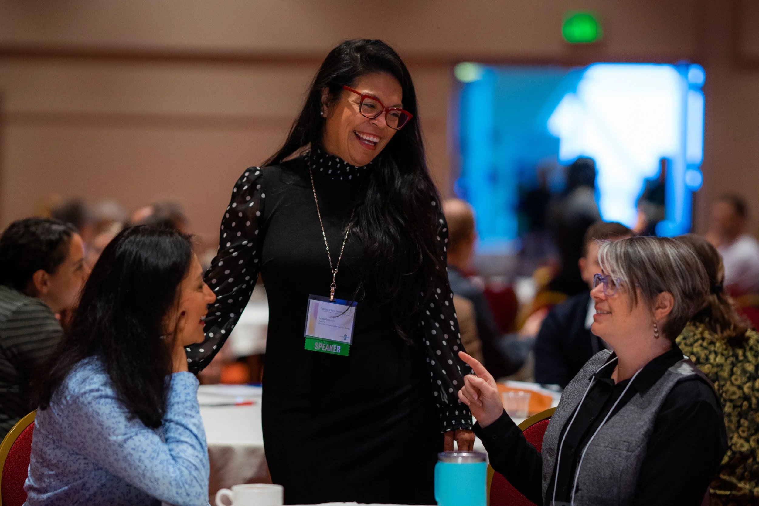 A woman speaking at a conference, smiling and engaging with seated attendees at a round table in a conference room.
