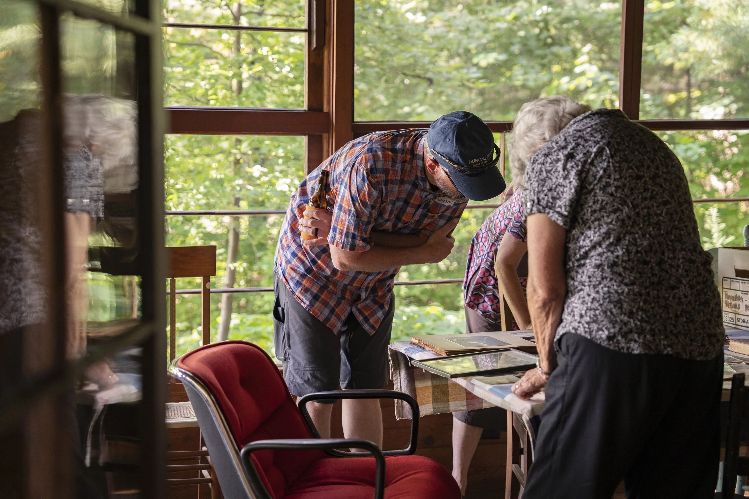 Three people, two women and one man, are gathered around a table inside a room with large windows showing a green, leafy outdoor scene. They are looking at papers or books on the table, leaning over and engaging in conversation.