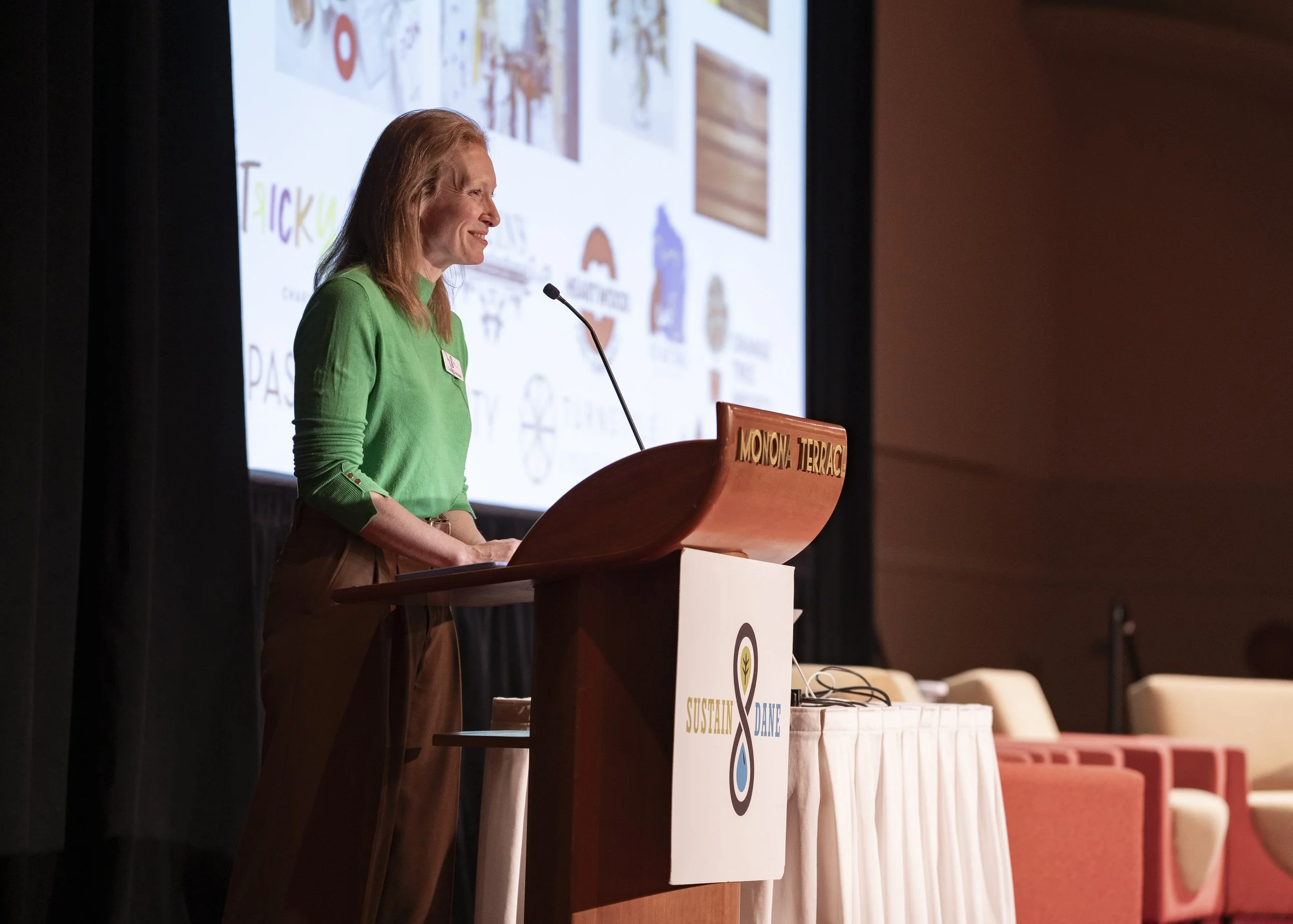 A woman with red hair wearing a green top stands at a wooden podium on stage, delivering a speech. Behind her, a large presentation screen displays various images and graphics. The podium has a sign that reads 'Moona Terrace' and a logo with the word