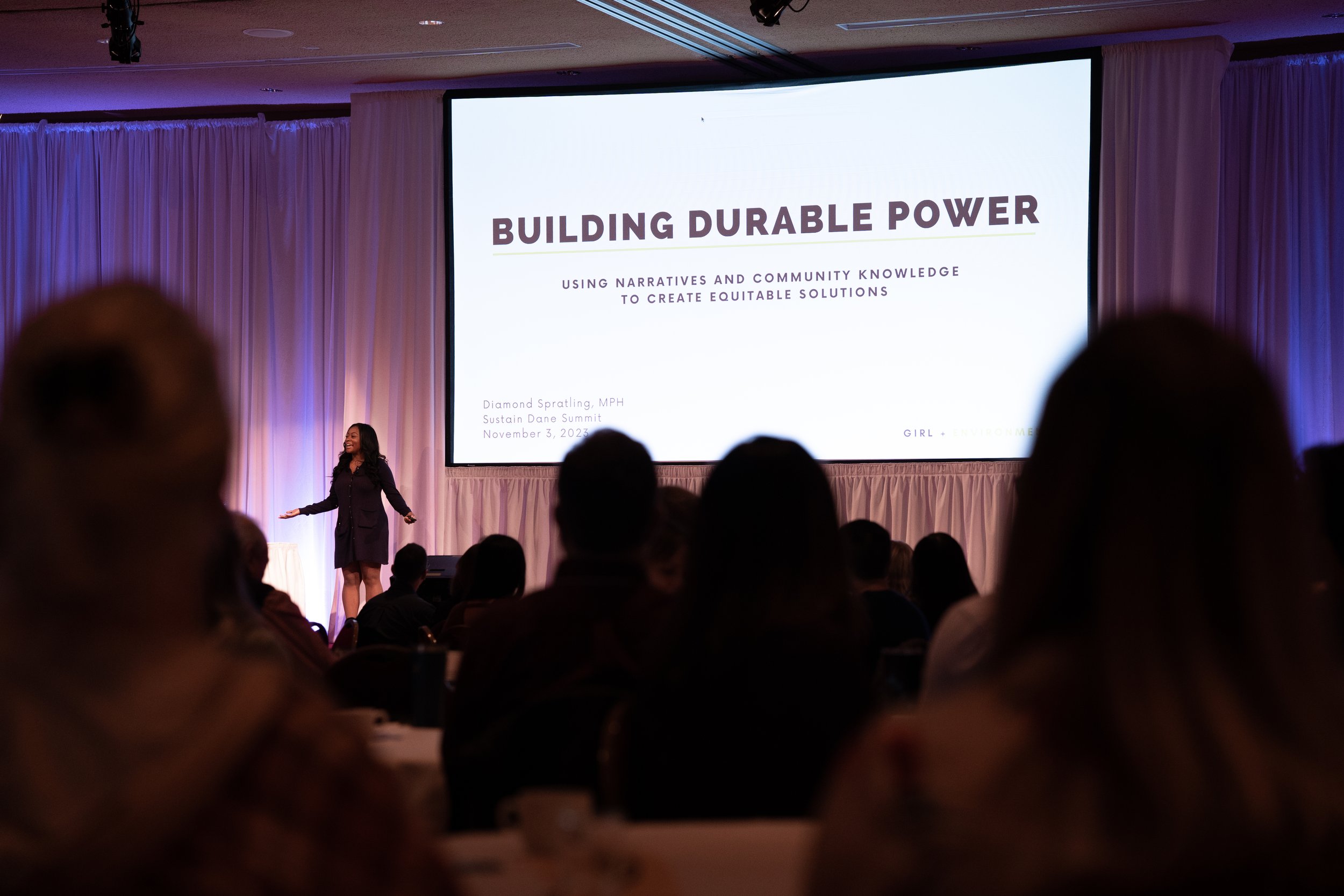 A woman standing on stage giving a presentation in front of a large screen that reads 'Building Durable Power' during a conference. The audience is seated and listening.