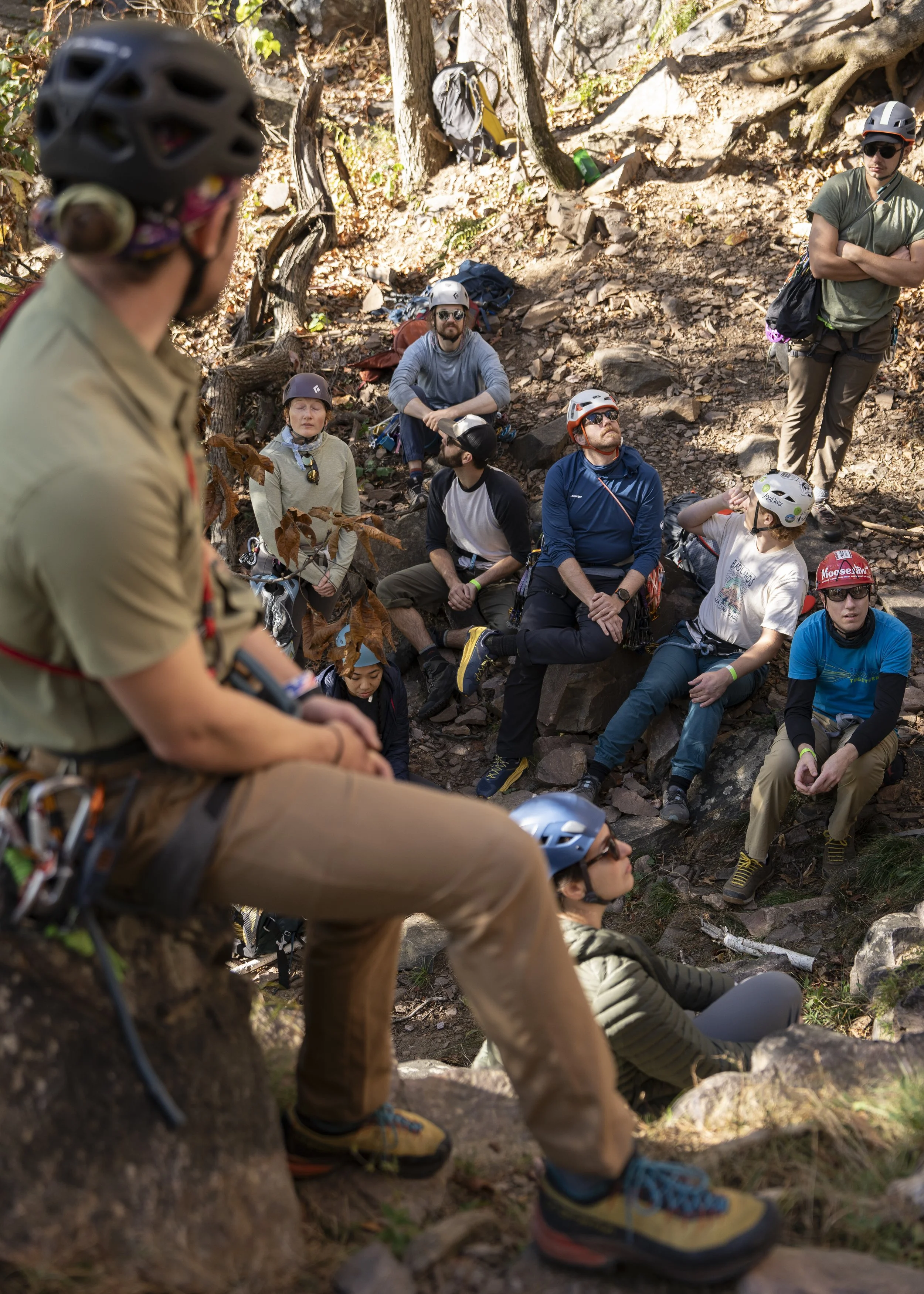 Group of people wearing helmets and outdoor clothing sitting and standing on a rocky hillside in a wooded area, listening to a person in the foreground.
