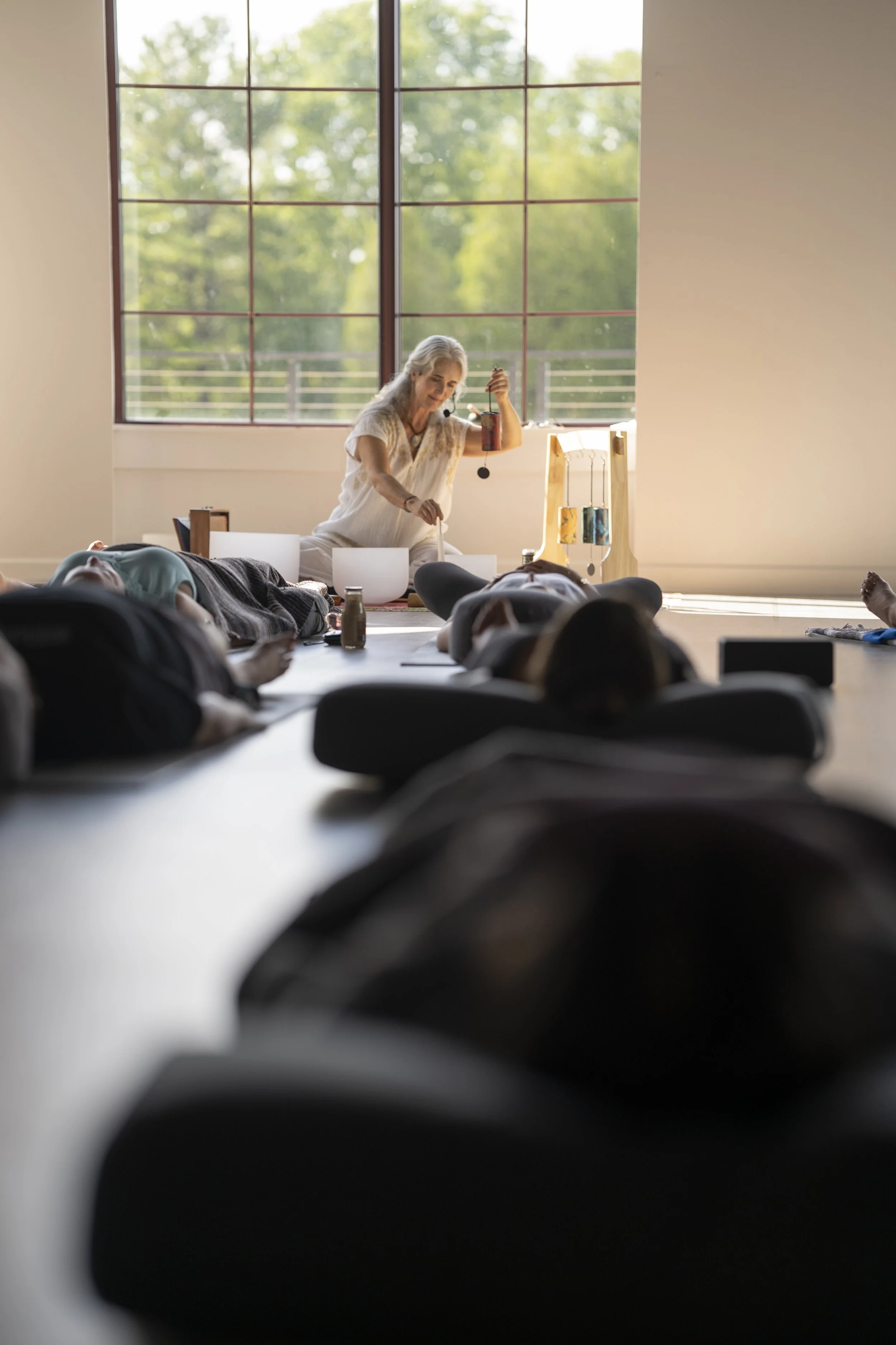 A woman conducting a sound healing session with multiple people lying on the floor, and sunlight streaming through large windows in the background.
