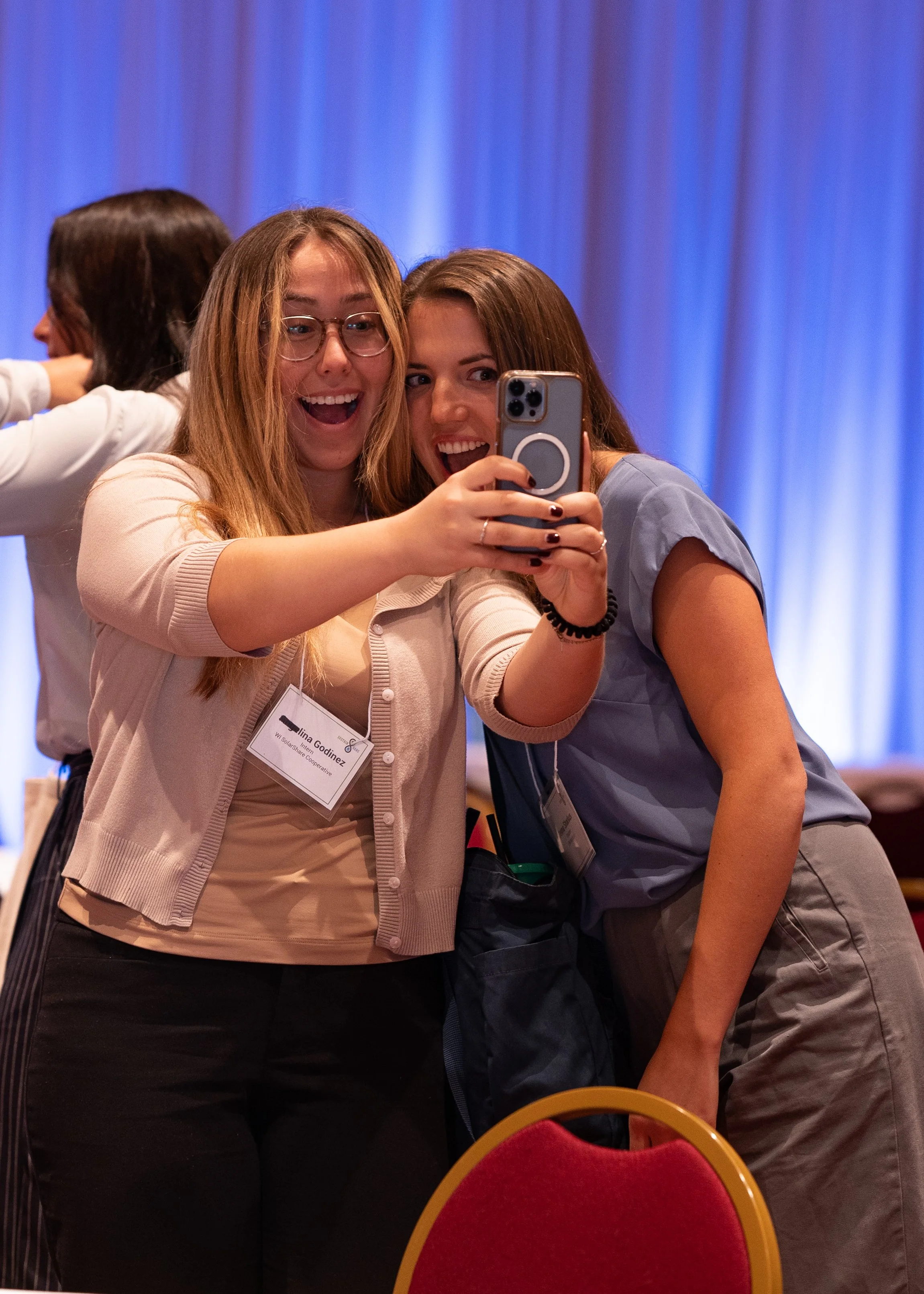Two young women taking a selfie at a conference. They are smiling and posing, with a blue curtain background and conference name tags.