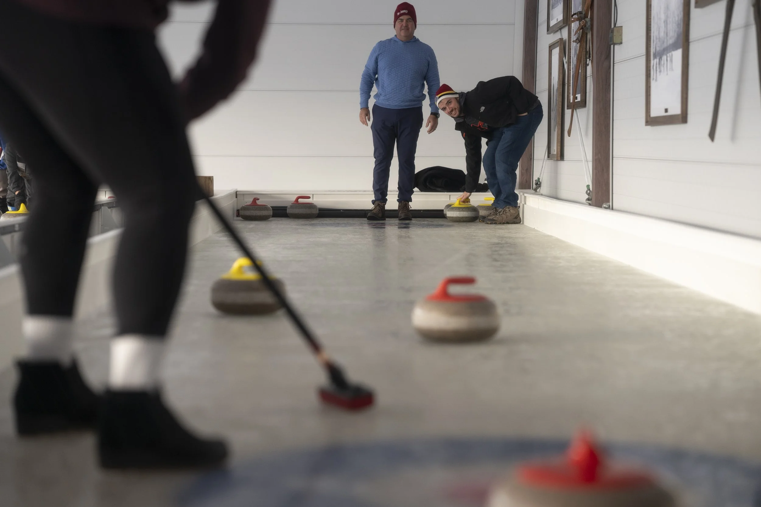 Two men are playing a game of indoor curling. One man is bent over, releasing a curling stone, while two other men are watching from the background.