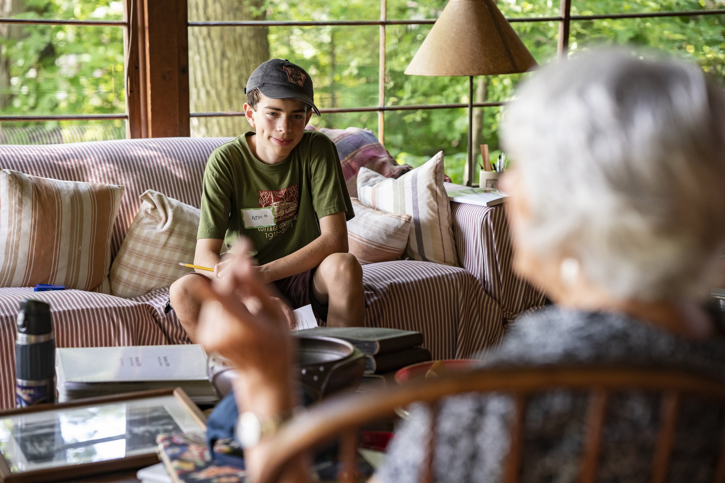 A young boy sitting on a striped couch, talking to an older woman with gray hair, in a room with large windows and green trees outside.