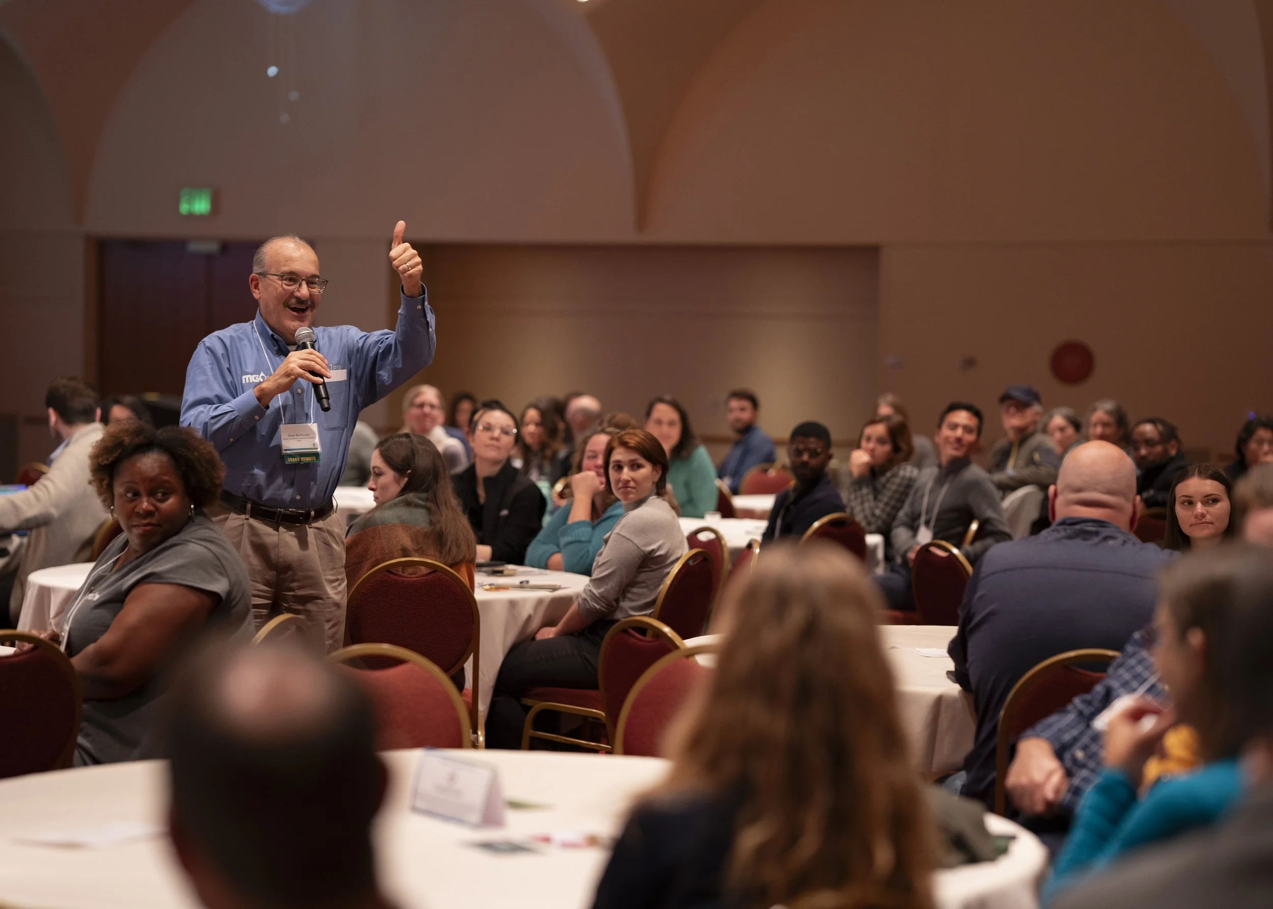 A man holding a microphone and giving a thumbs-up gesture while speaking to an audience in a conference room filled with seated attendees.