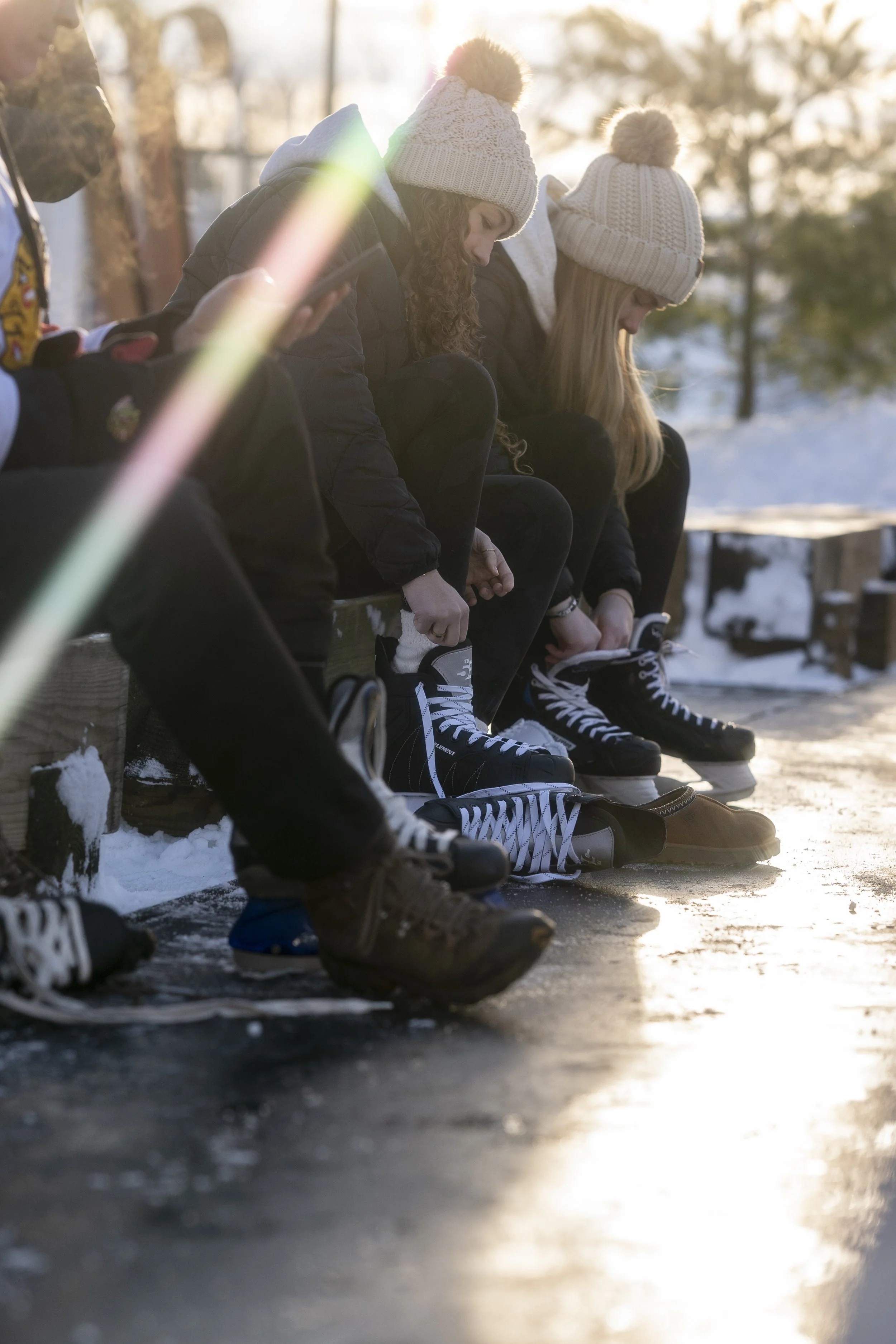 Three women sitting on a bench outdoors in winter, putting on ice skates, with snow and trees in the background and sunlight shining behind them.