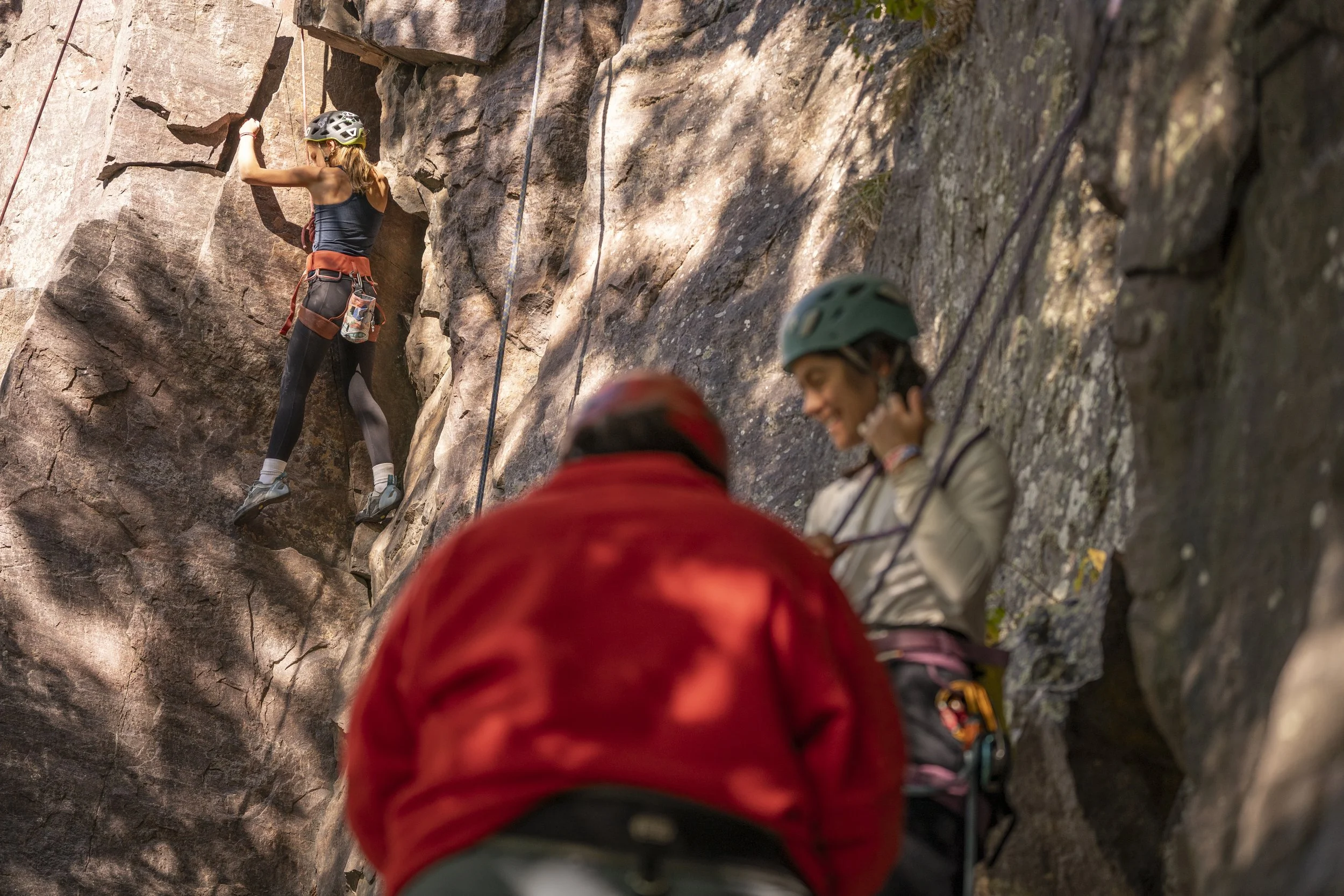 A woman in climbing gear ascending a rock wall with two people assisting below.